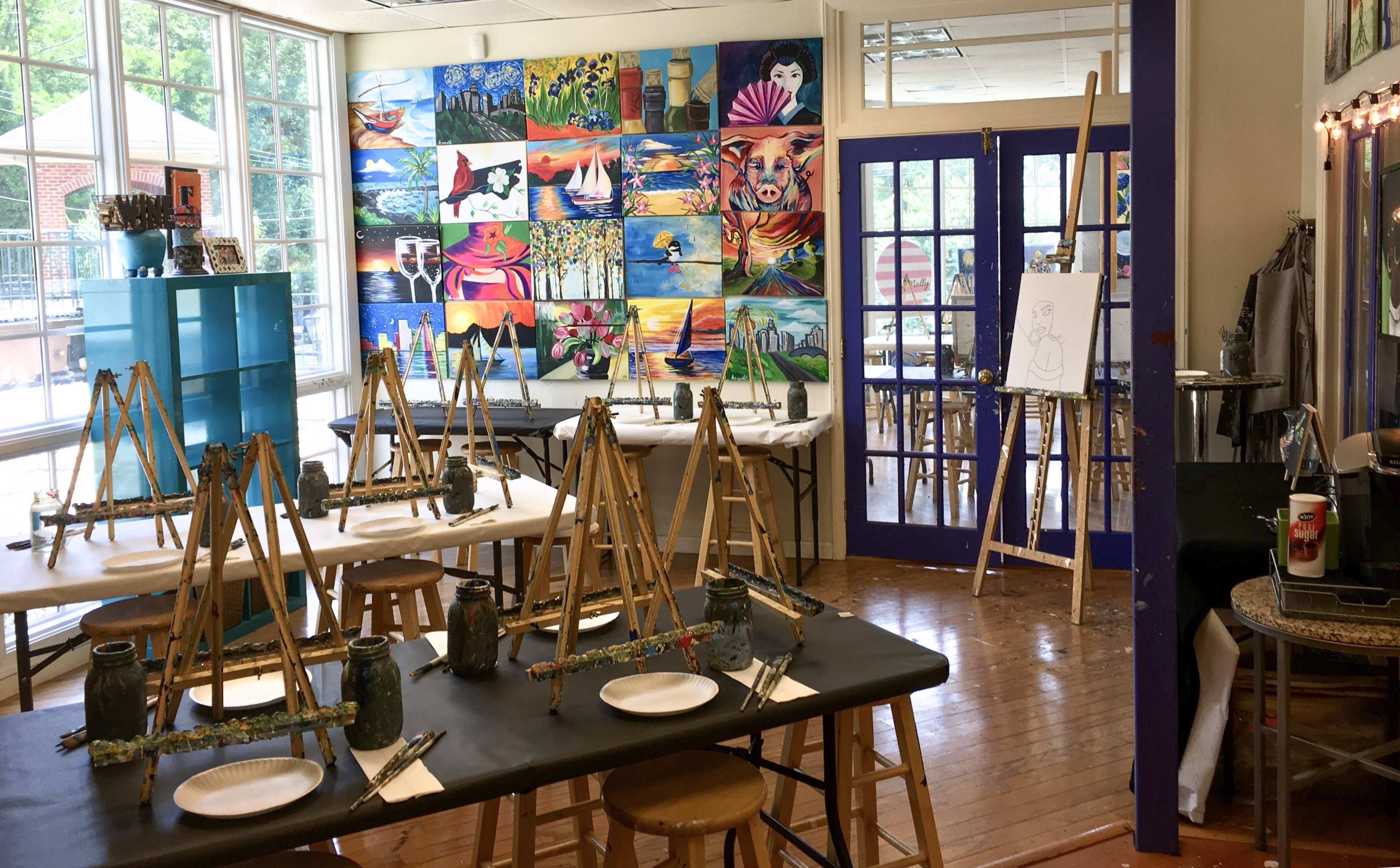 An art studio with several easels set up around tables and a colorful mural of paintings on the wall.