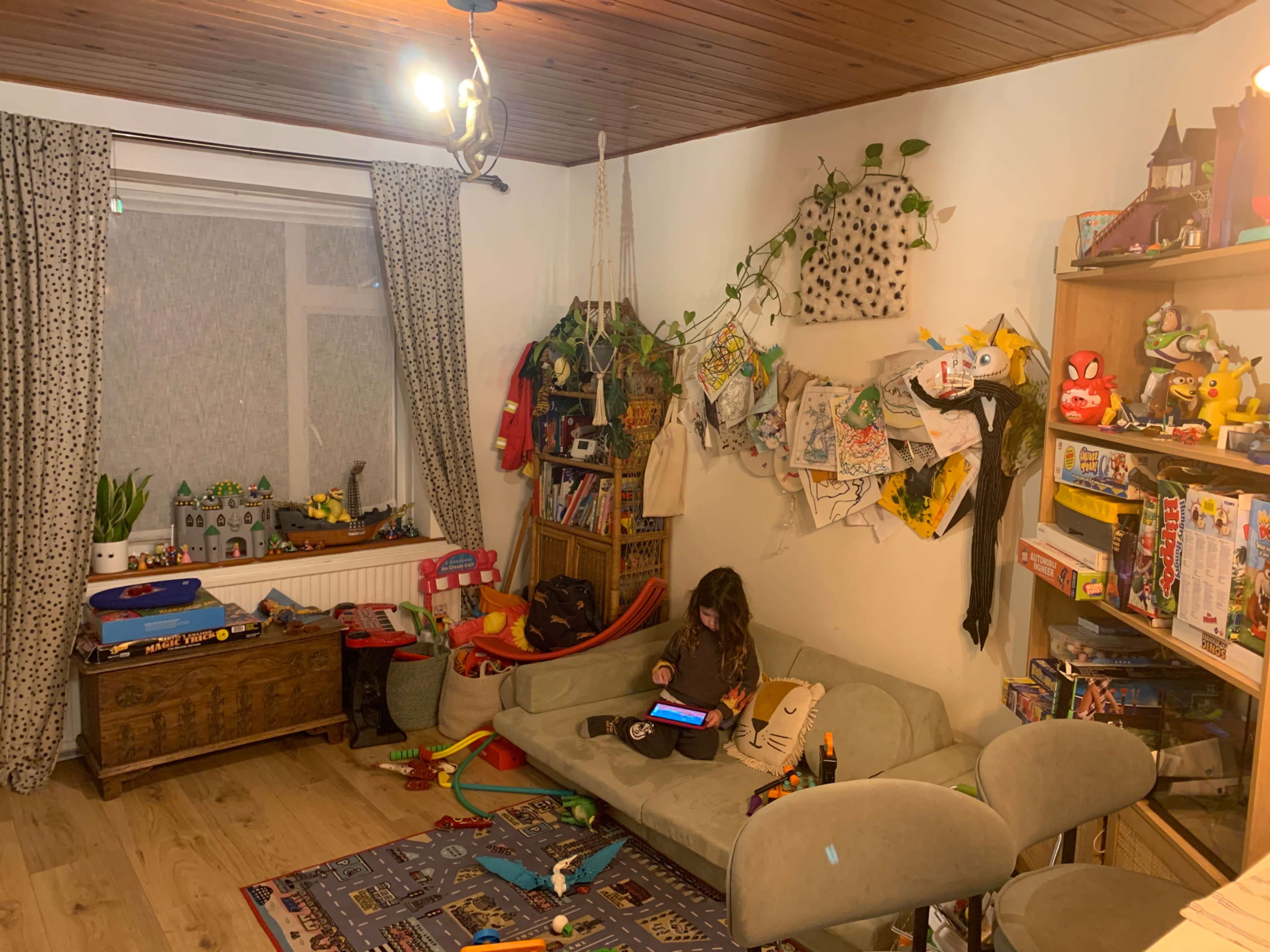 A child sits on a couch in a cluttered living room filled with toys, books, and artwork hanging on the walls.