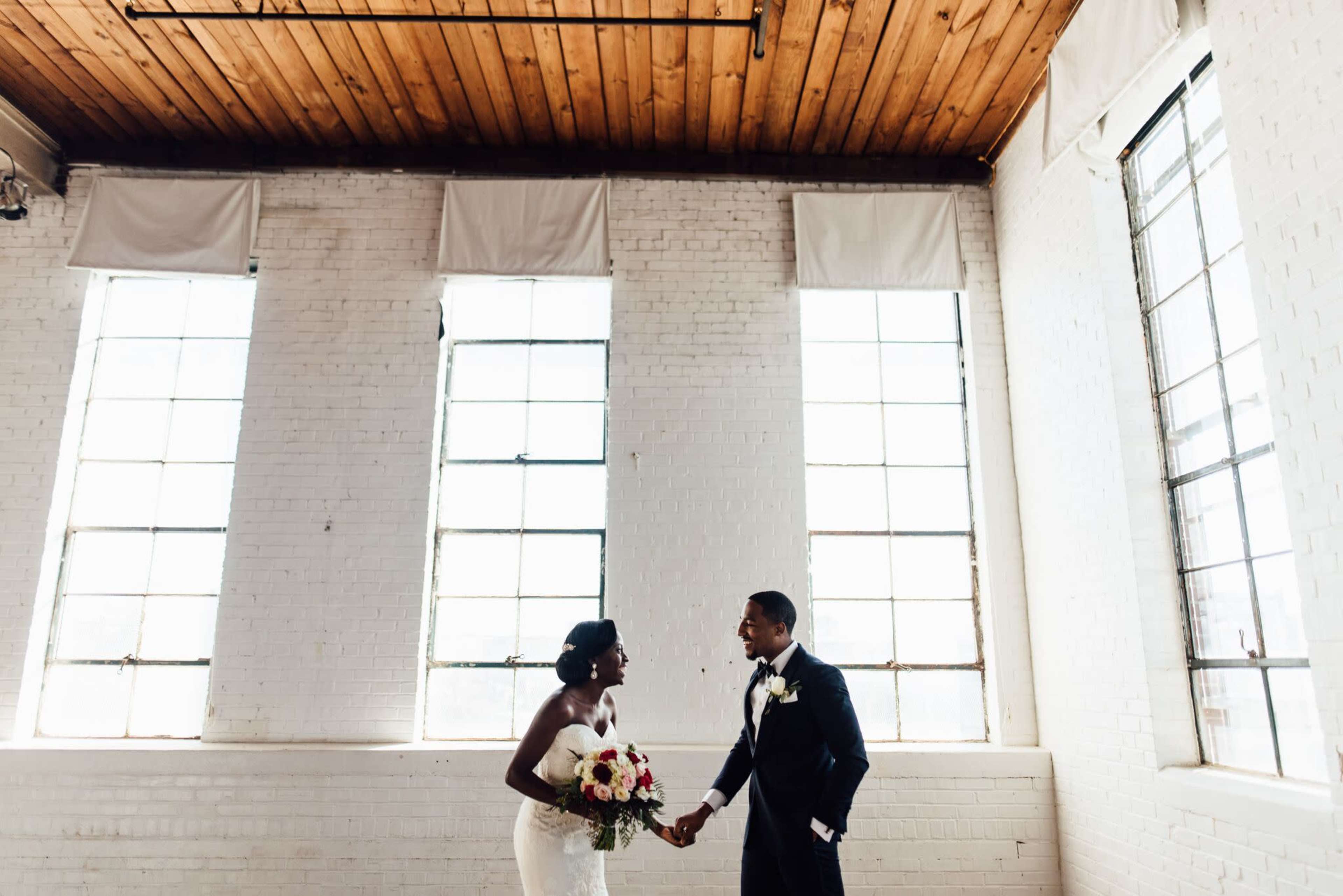 A bride and groom hold hands and smile at each other in a well-lit, airy room with large windows and exposed wood beams.