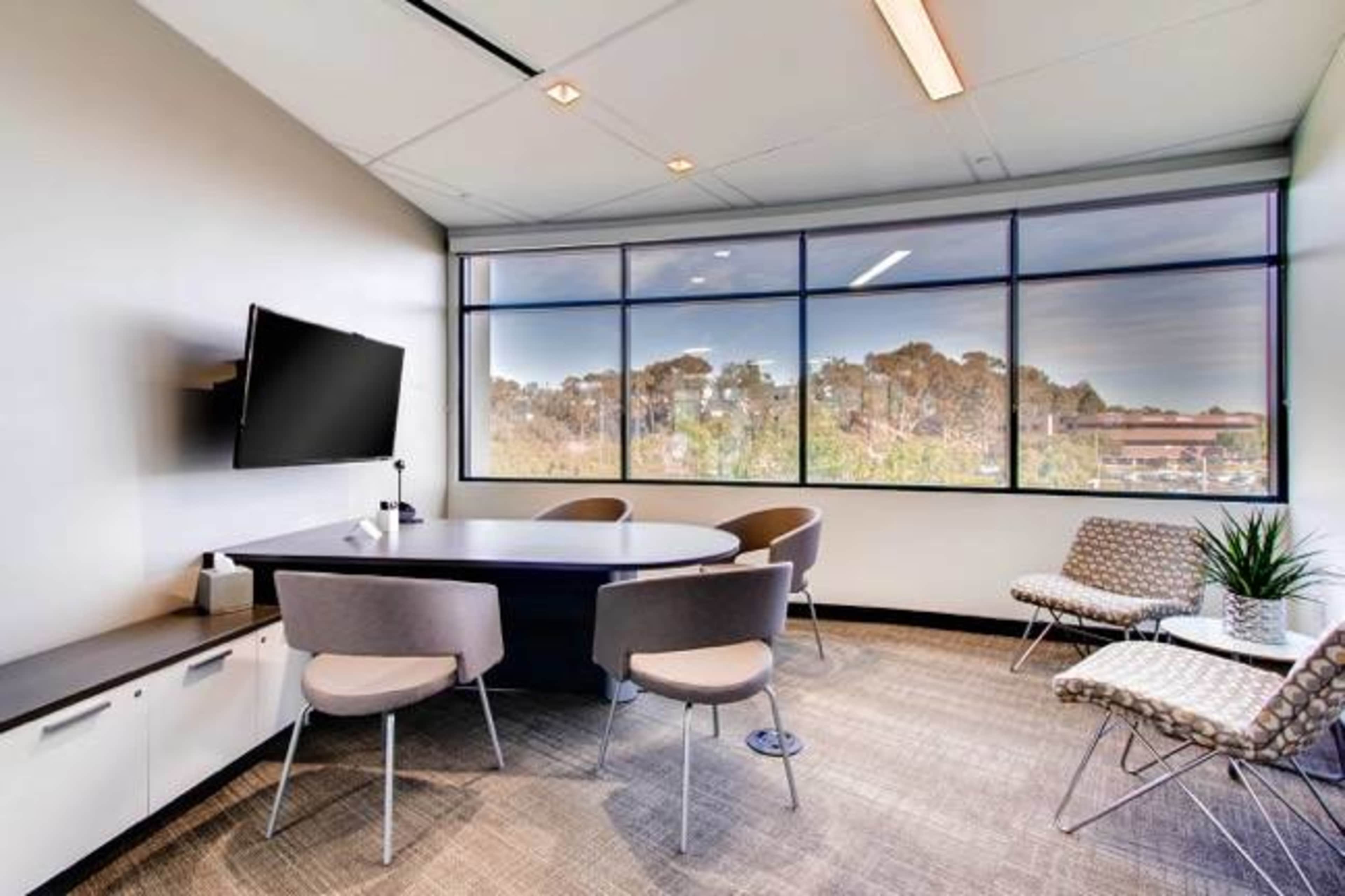 The image shows a modern meeting room with a round table, four chairs, a wall-mounted television, and large windows overlooking a green landscape.