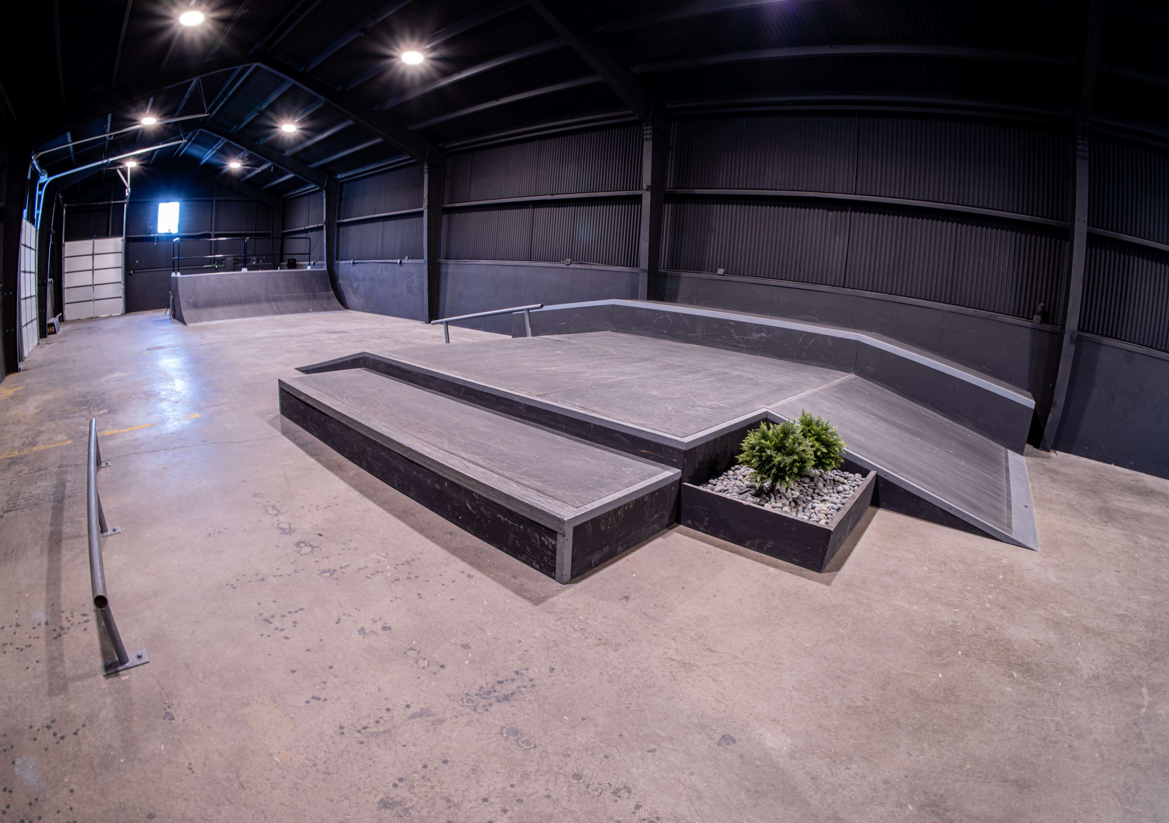 The image shows an indoor skateboarding facility featuring wooden ramps, a ledge, and a small planter in the center of the space.