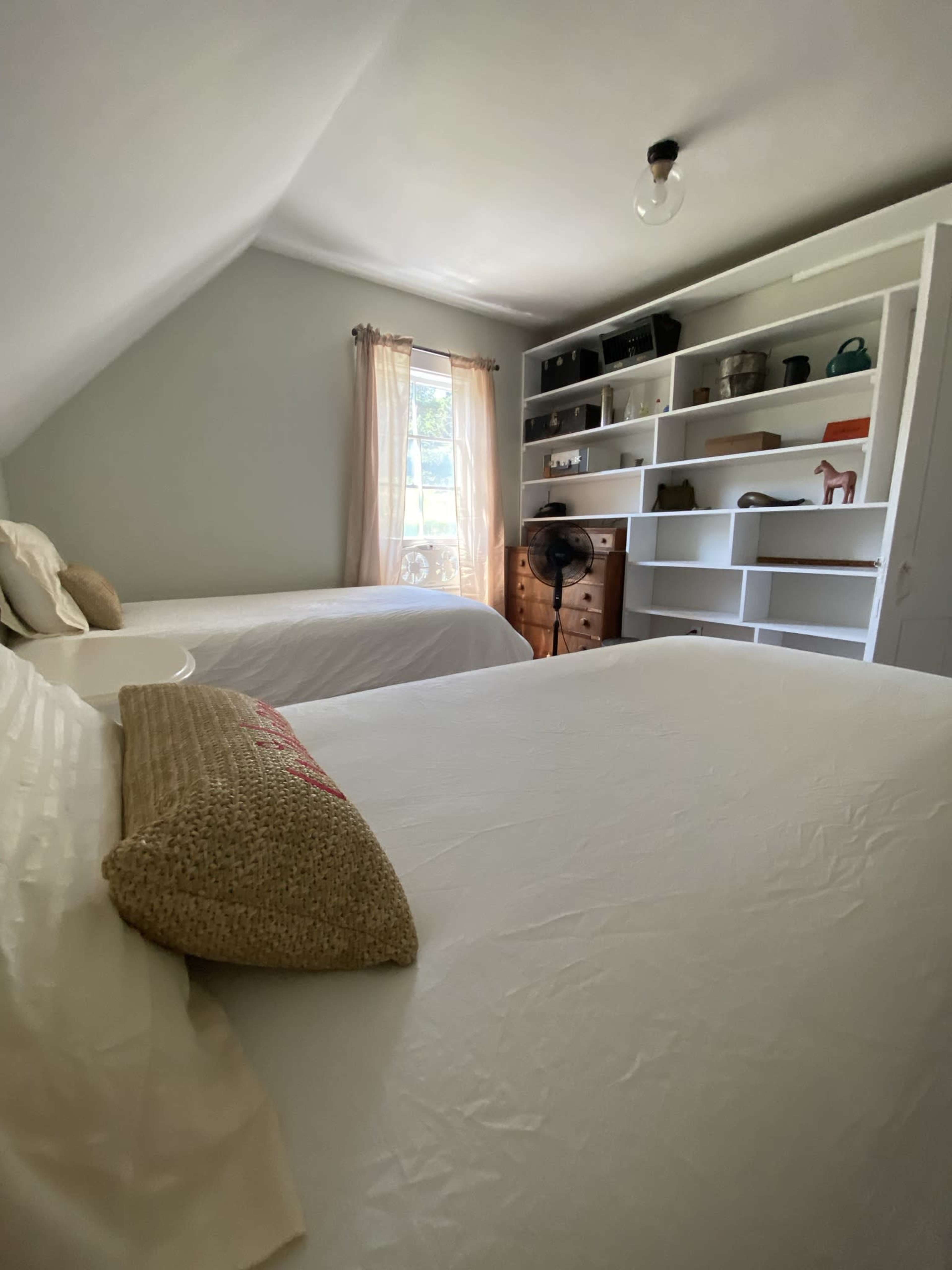 A tidy bedroom featuring two beds, a window with sheer curtains, and a bookshelf filled with various items.