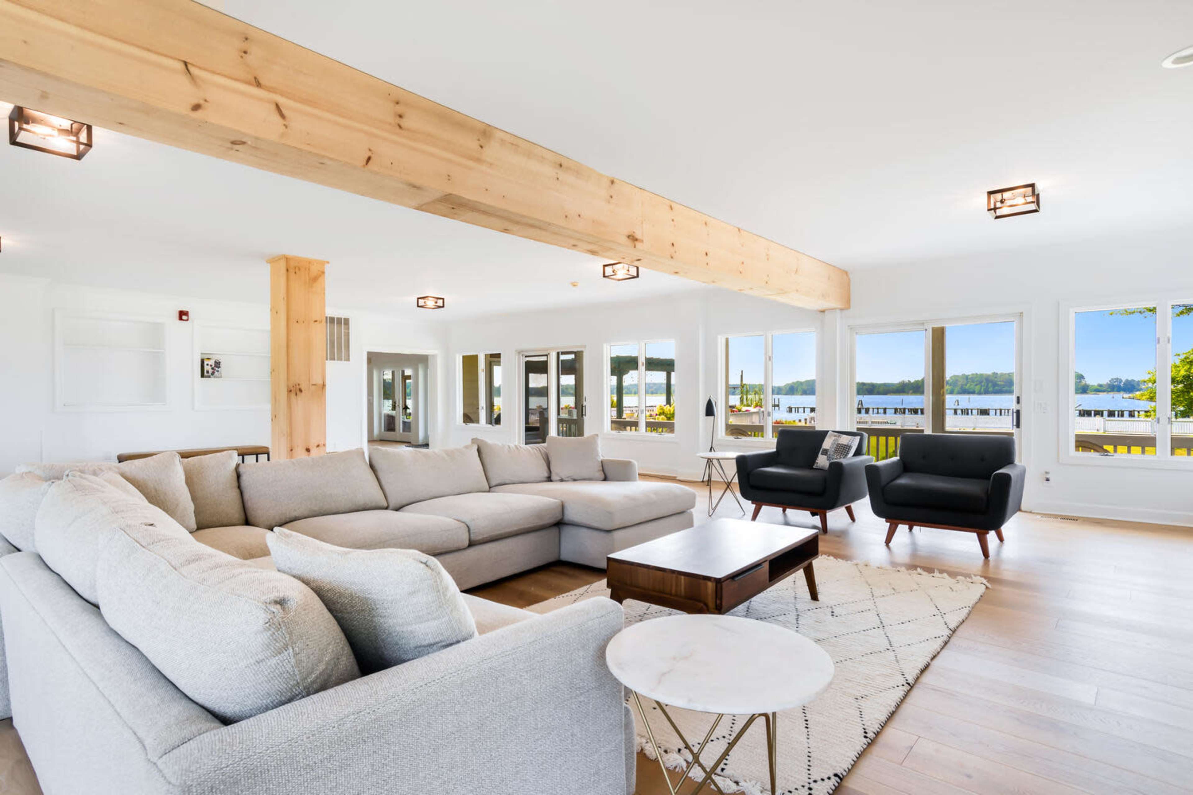 A modern living room with large windows, a spacious gray sectional sofa, two black armchairs, a wooden beam, and a coffee table on a light-colored rug.