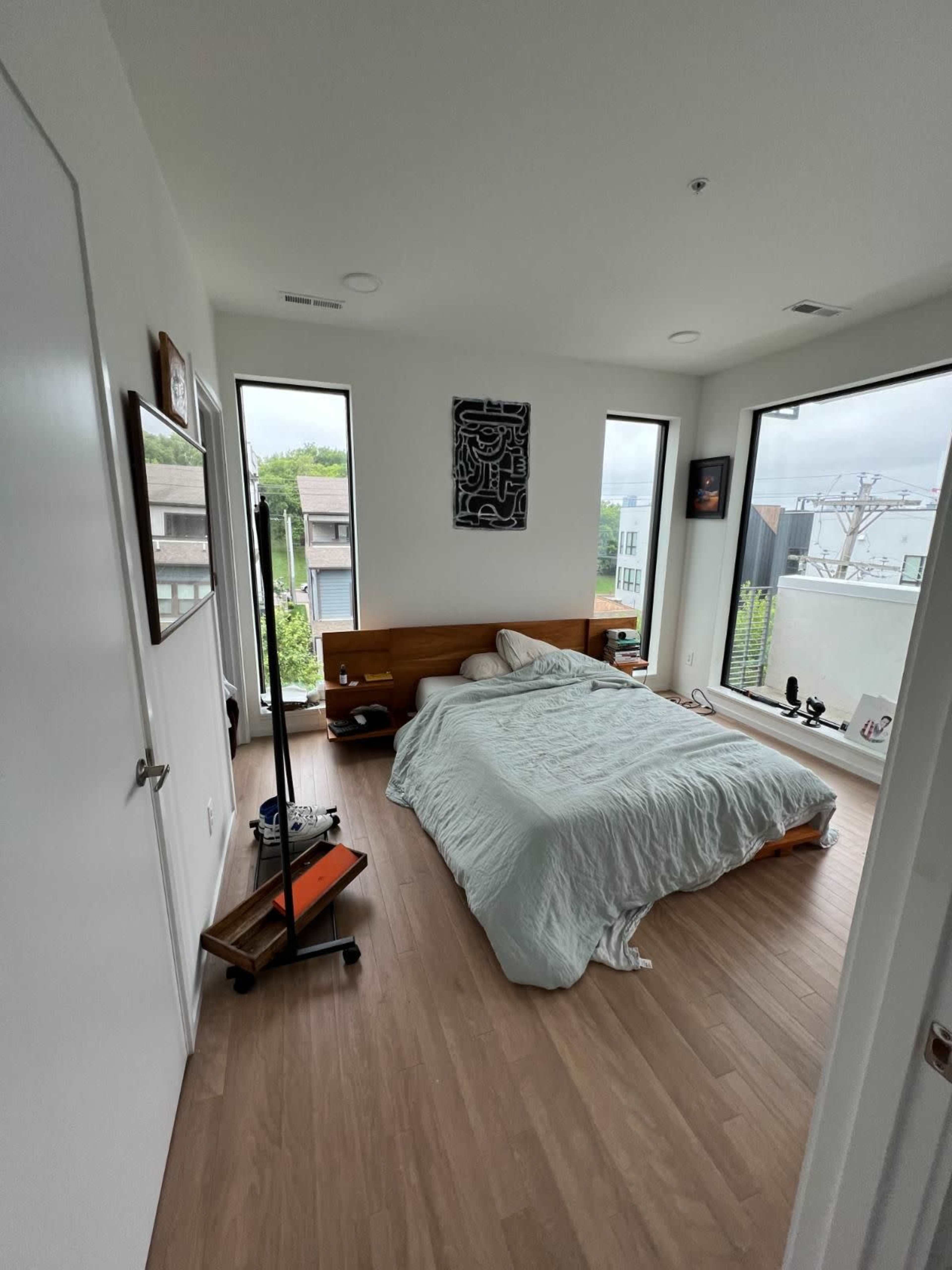 The bedroom features a wooden bed with a light-colored comforter, large windows, and minimal furniture, set in a modern architectural space.