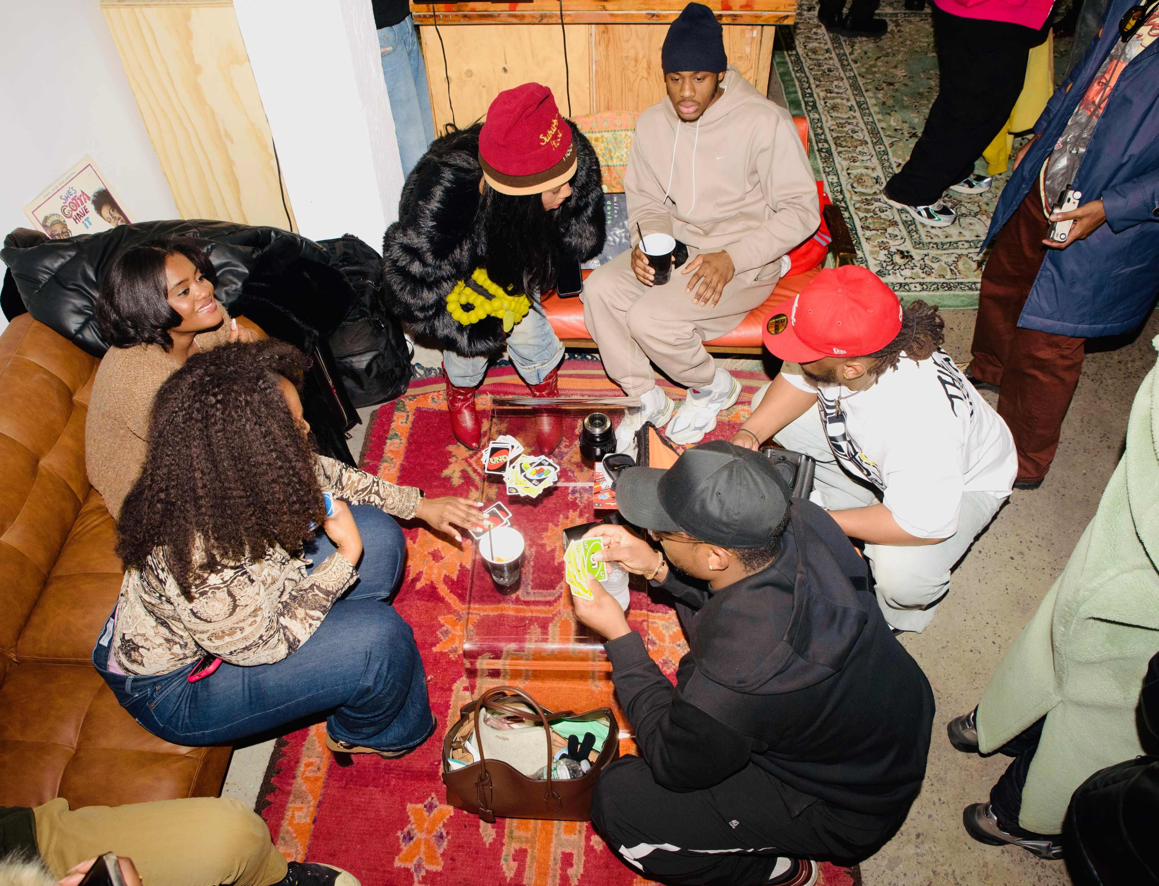 A group of people sits on a couch and floor, engaging in conversation and playing a game on a colorful rug in a lively indoor space.