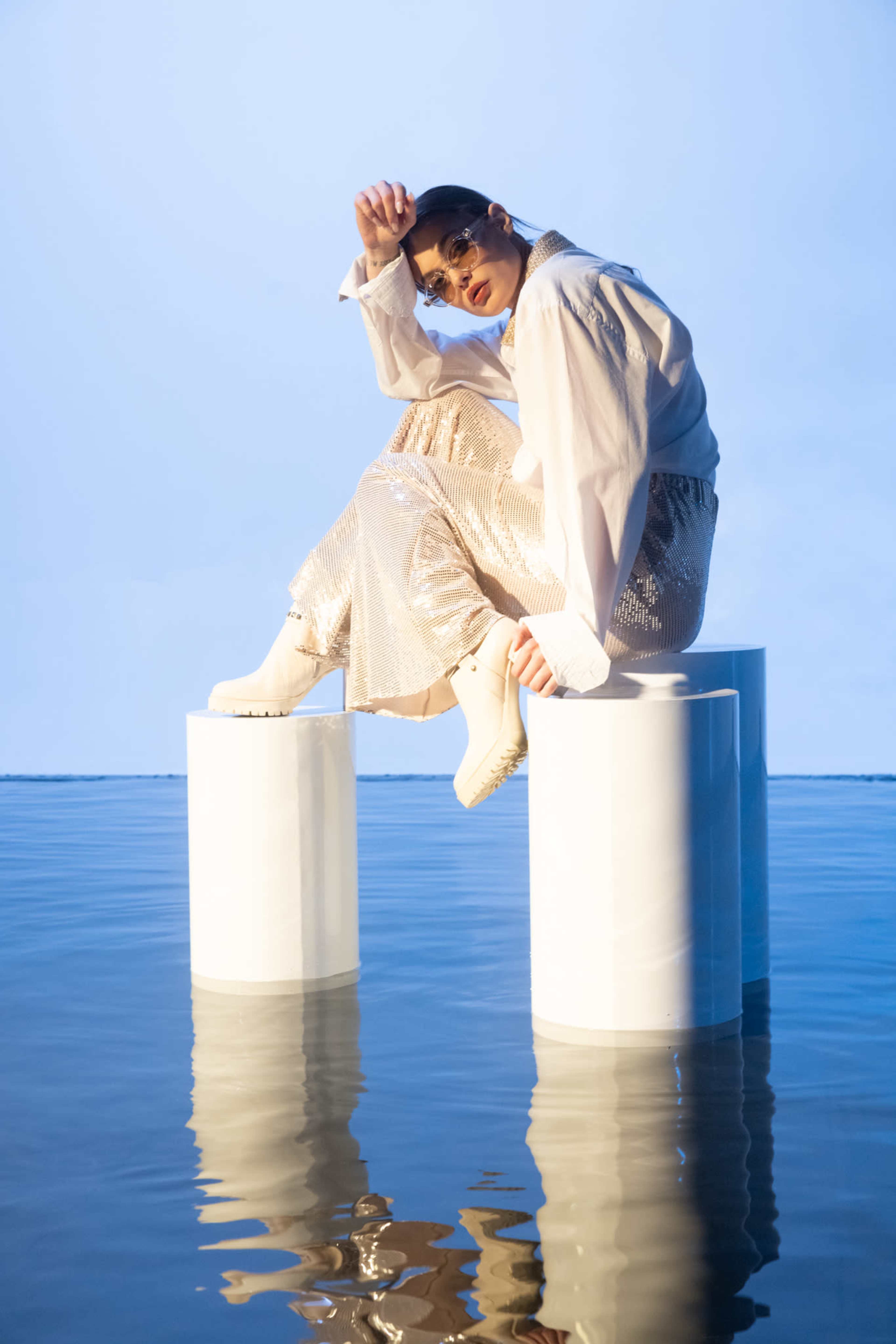 A person wearing a white shirt and shiny skirt sits on a white cylindrical pedestal in water, posing against a blue sky.