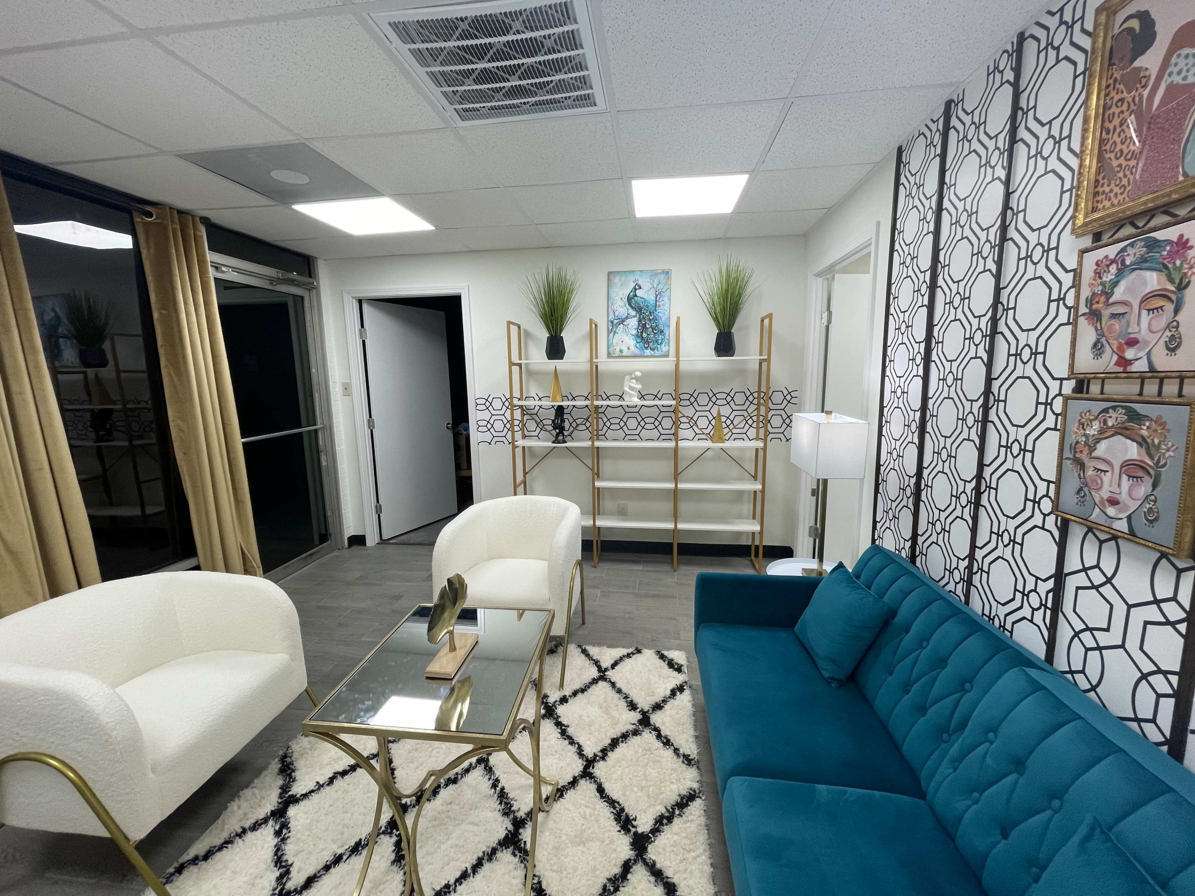 The image shows a modern lounge area with a teal sofa, two white armchairs, a glass coffee table, and decorative wall art, featuring a shelving unit and plants.