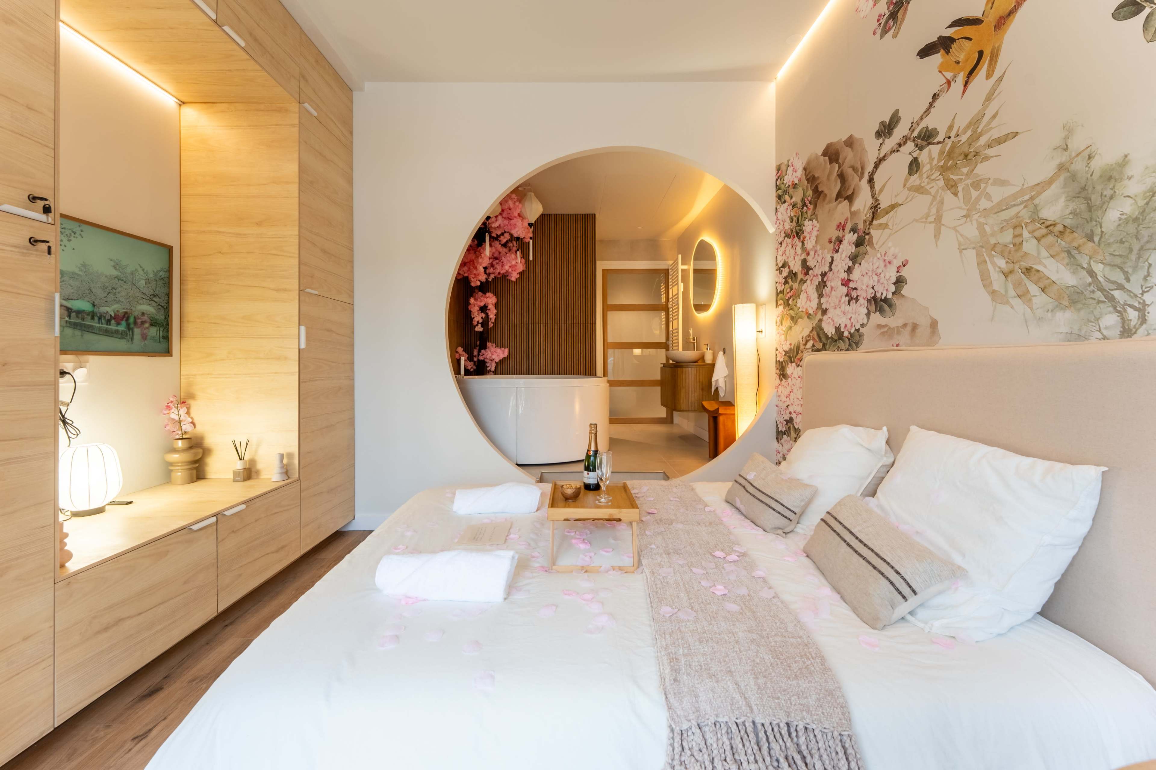 A modern bedroom features a circular cutout leading to a bathroom, with floral wall art and a neatly made bed adorned with soft linens.