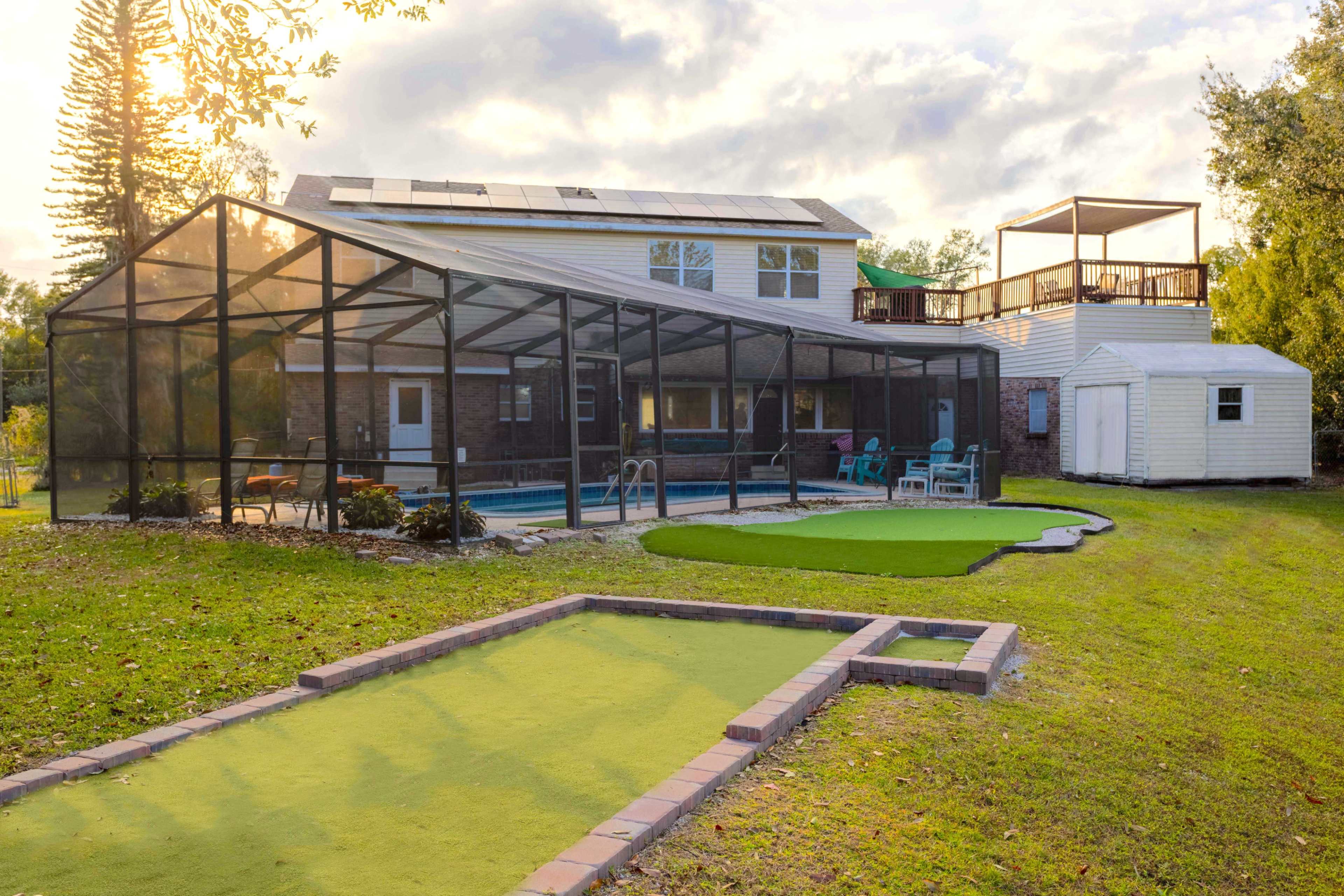 The image shows a residential backyard featuring a screened-in pool area, a small putting green, and a two-story house with a rooftop deck.