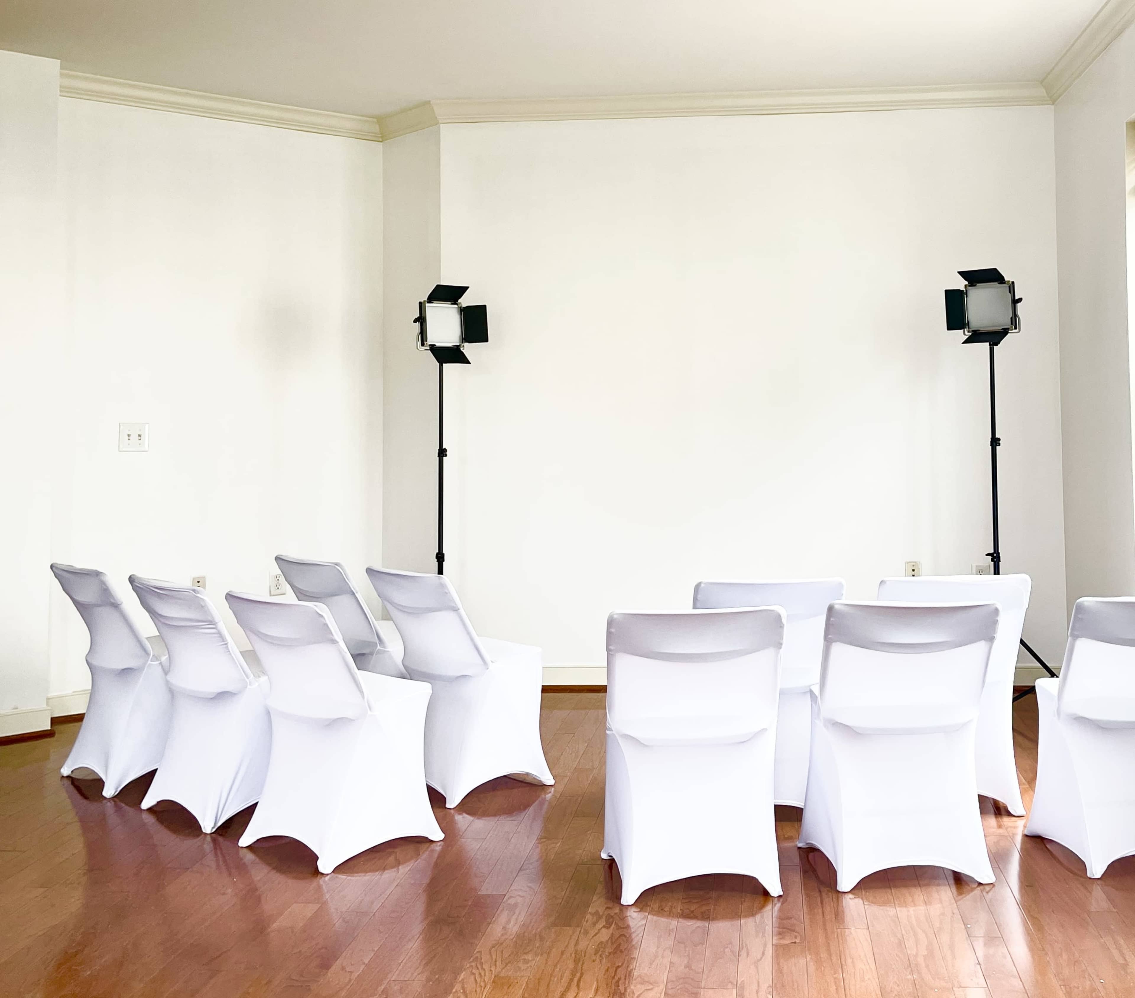 A spacious, well-lit room features several white chairs arranged in rows facing a blank wall, with two lighting fixtures positioned on either side.