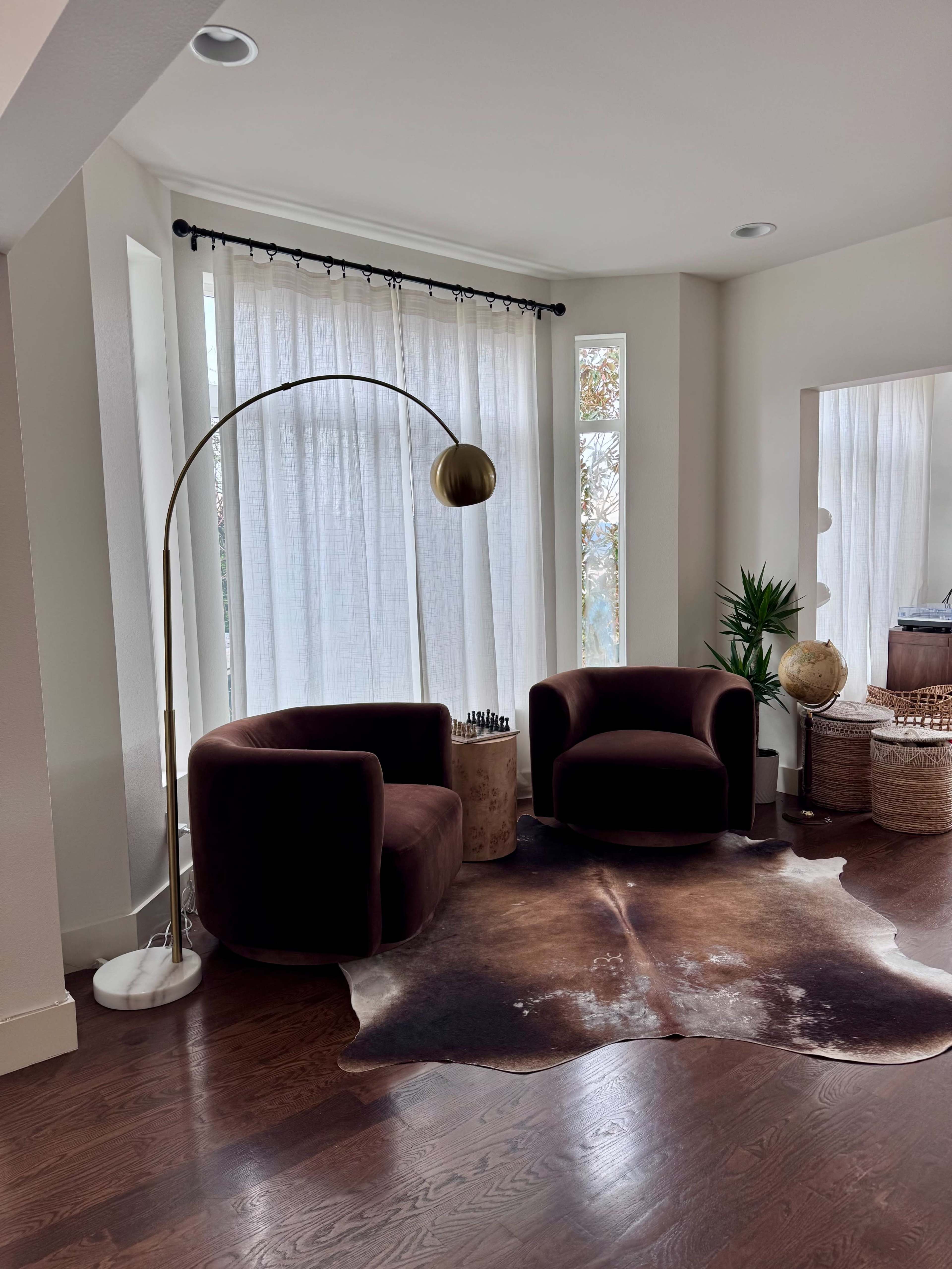 The image shows a cozy seating area featuring two brown velvet chairs, a standing lamp, a small round table, and a large window with sheer white curtains.