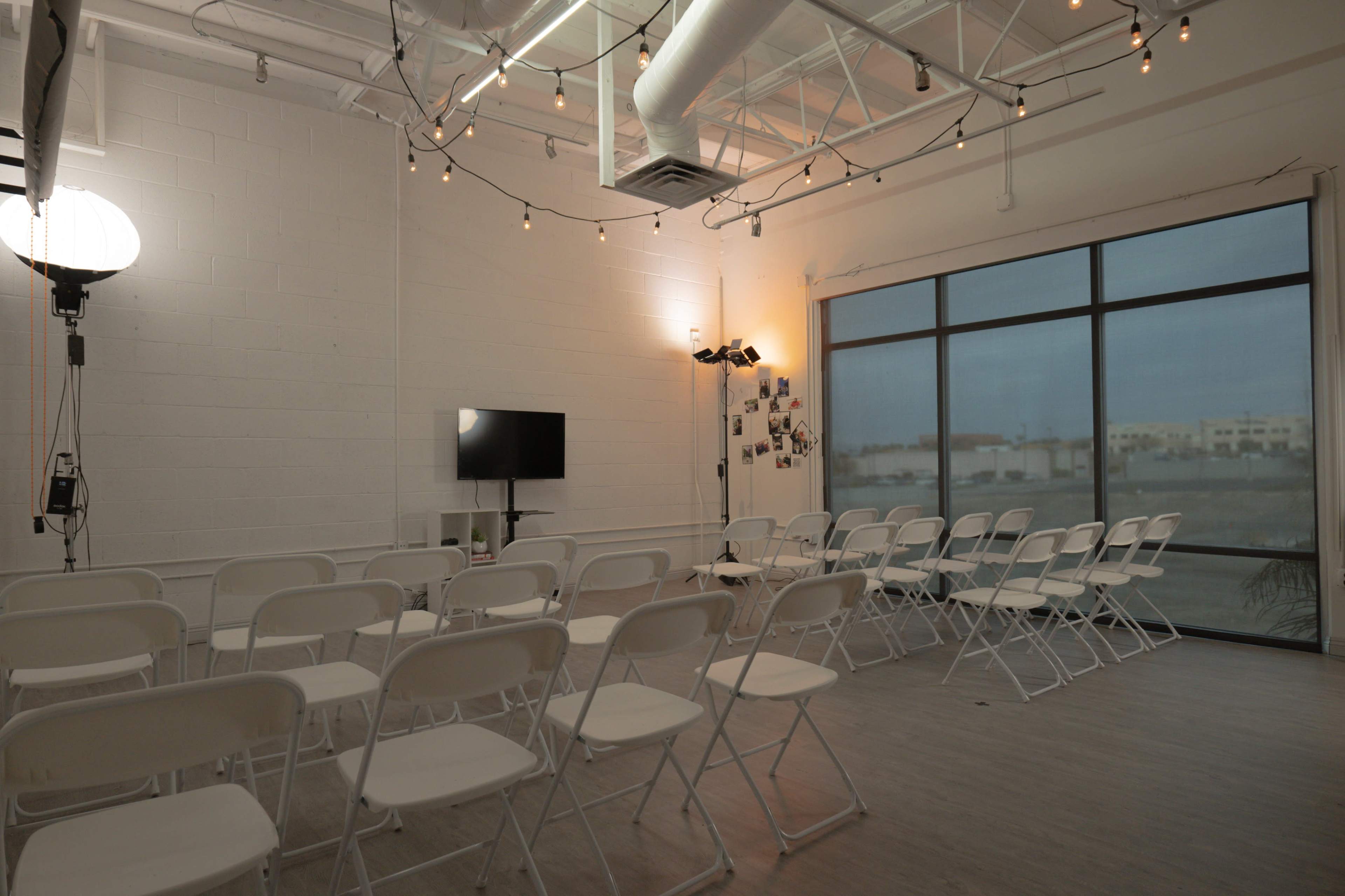 A spacious room with white walls, featuring a row of white folding chairs facing a television on a stand, with string lights hanging from the ceiling.