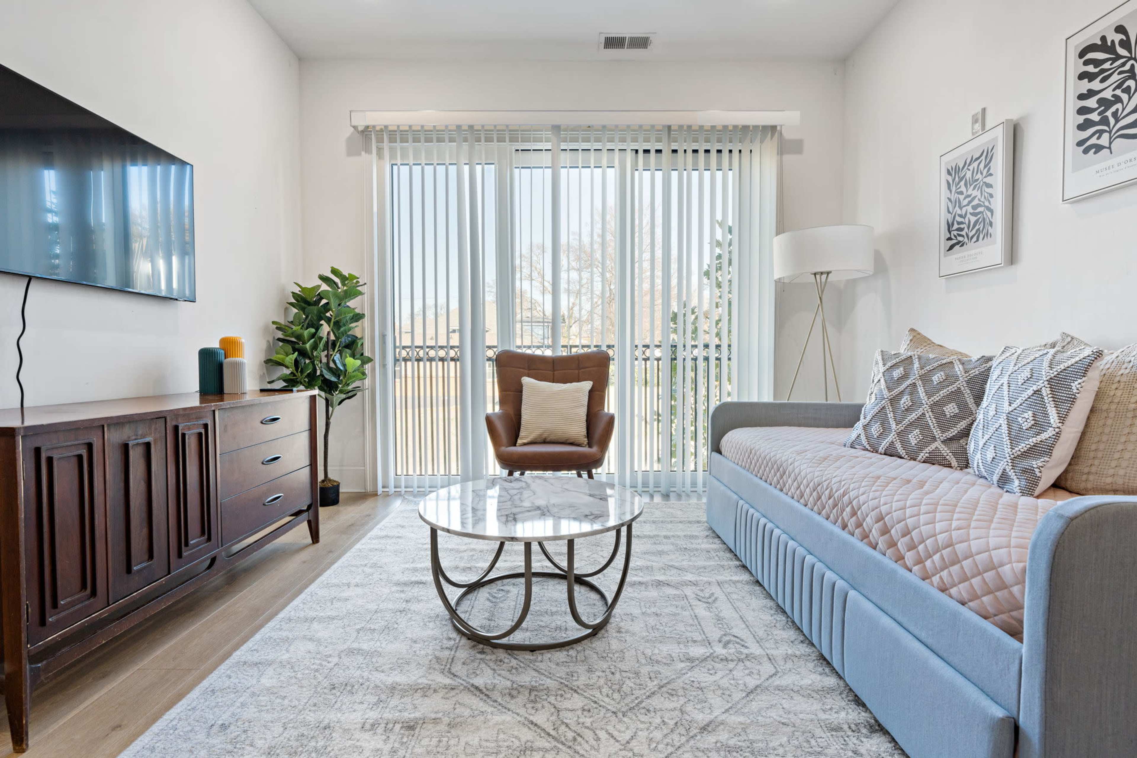 A modern living room features a sofa, a round coffee table, a television mounted on the wall, and large sliding glass doors with vertical blinds.