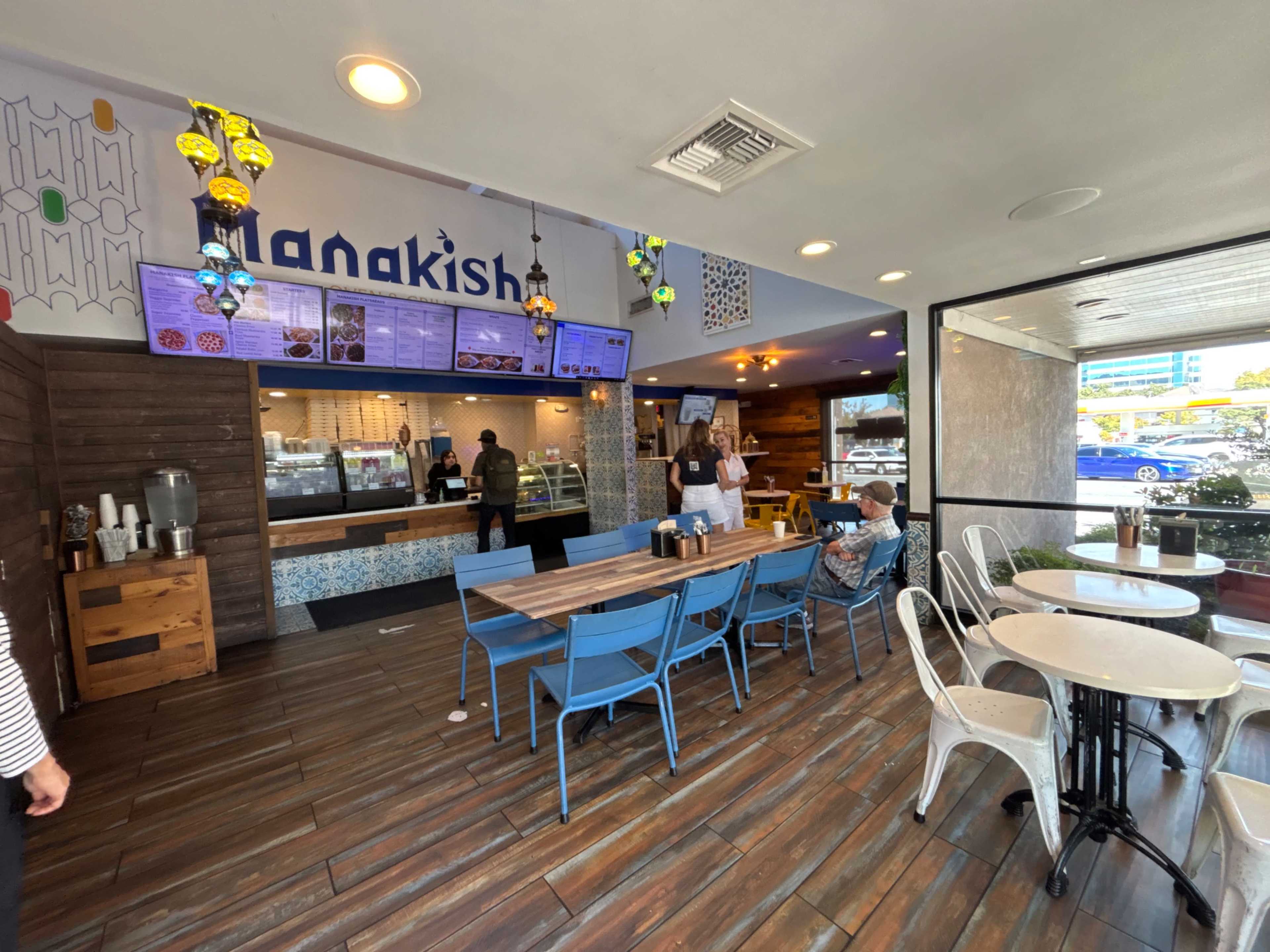 The image shows a modern eatery with a counter offering Lebanese food, featuring colorful hanging lights and a combination of blue and white seating arrangements.