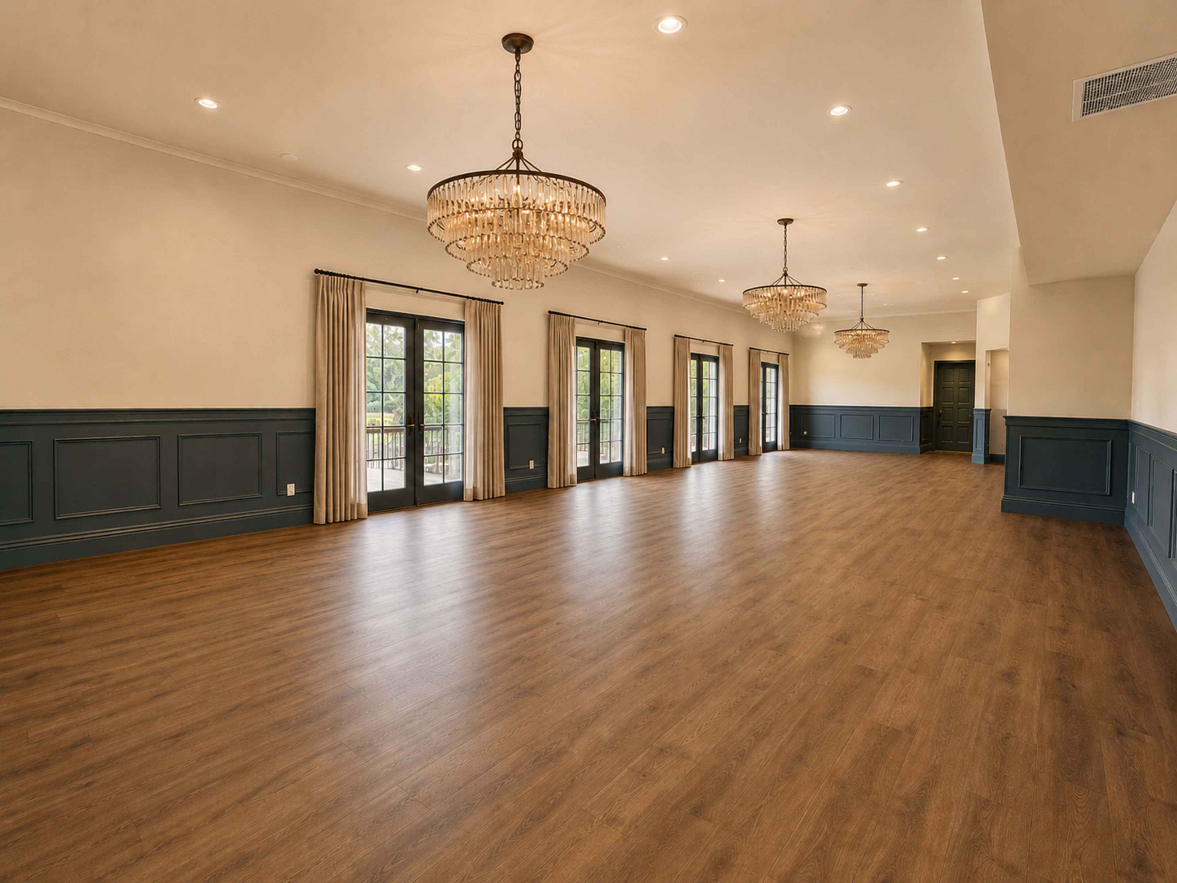 The spacious interior features wooden flooring, large French doors, and two ornate chandeliers hanging from the ceiling.