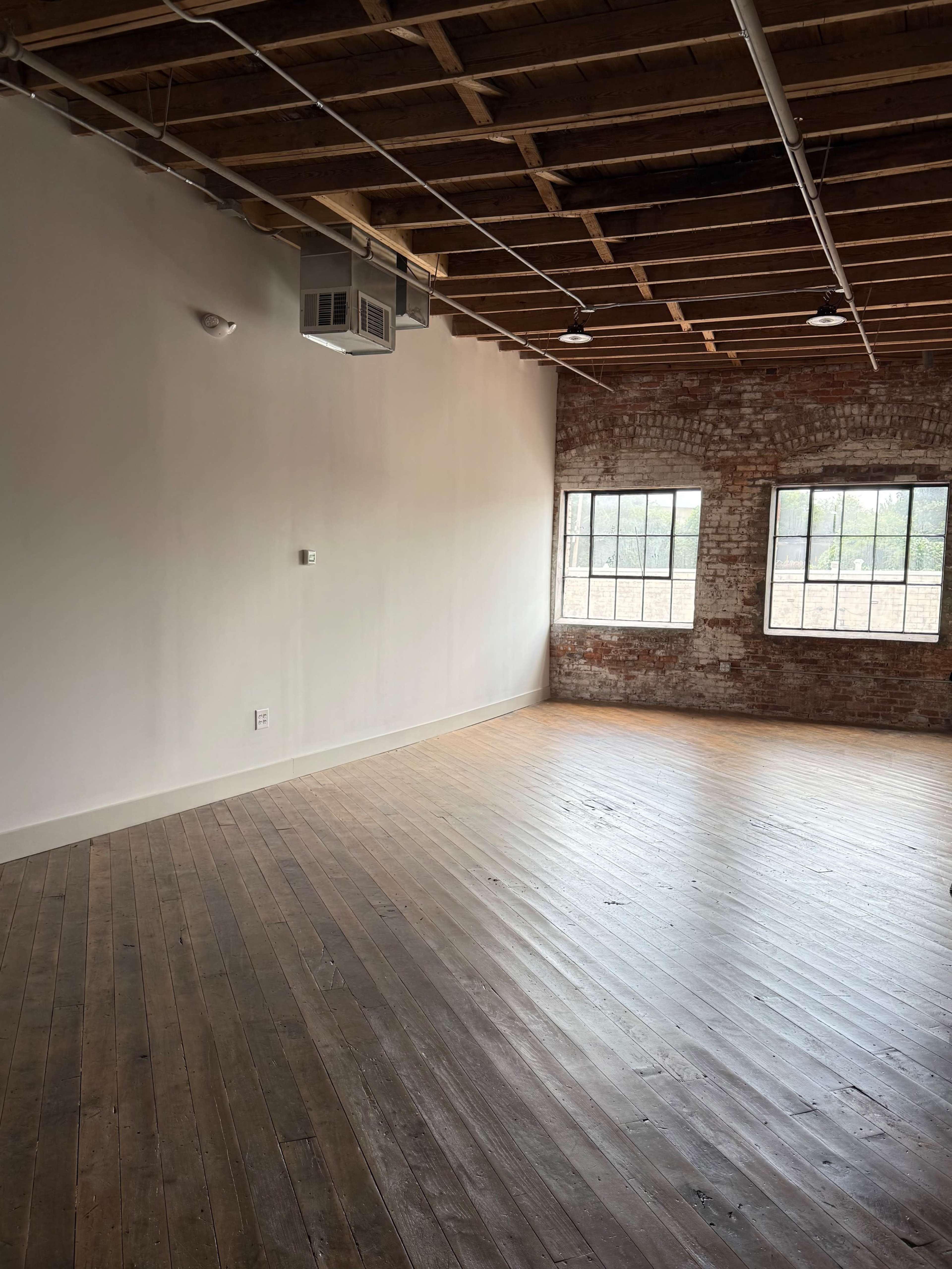 The image shows an empty room with exposed brick walls and wooden floors, featuring two large windows and a ceiling with visible beams.