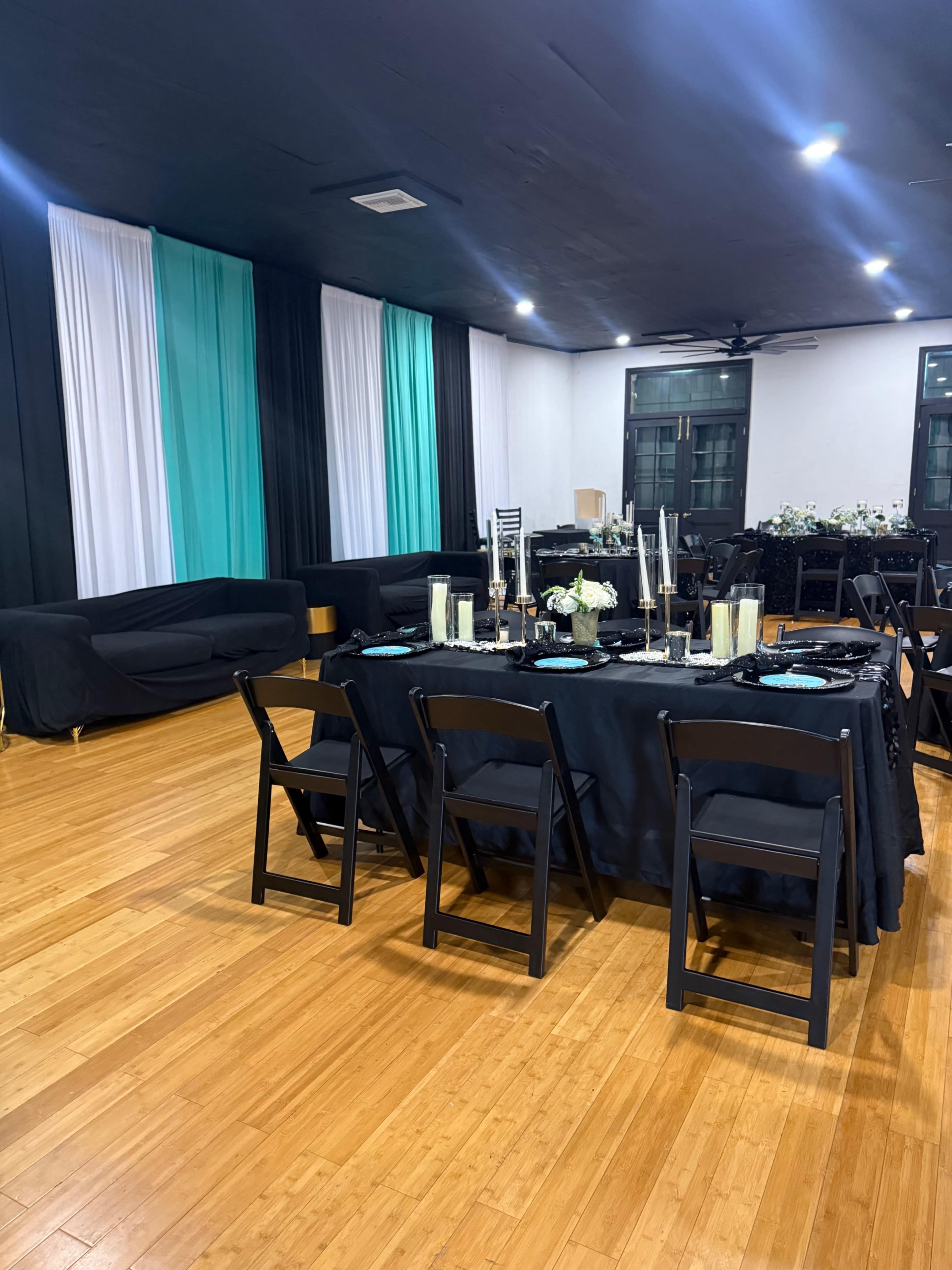 A large dining area features a black table set with candles and decorations, surrounded by black folding chairs, with teal and black drapery along the walls.