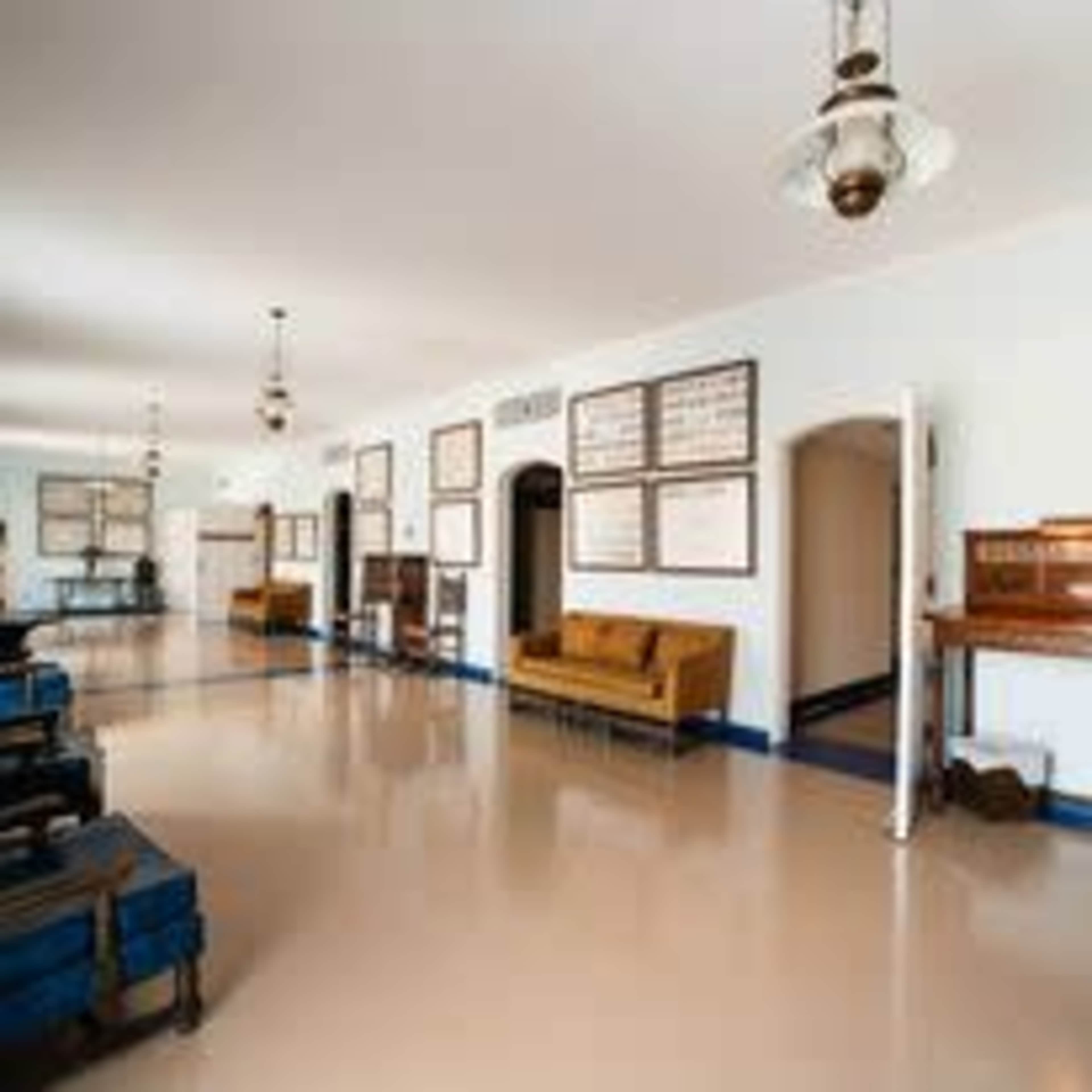 A long, well-lit hallway with beige walls, hanging light fixtures, and a row of vintage furniture, including a brown sofa and a piano.