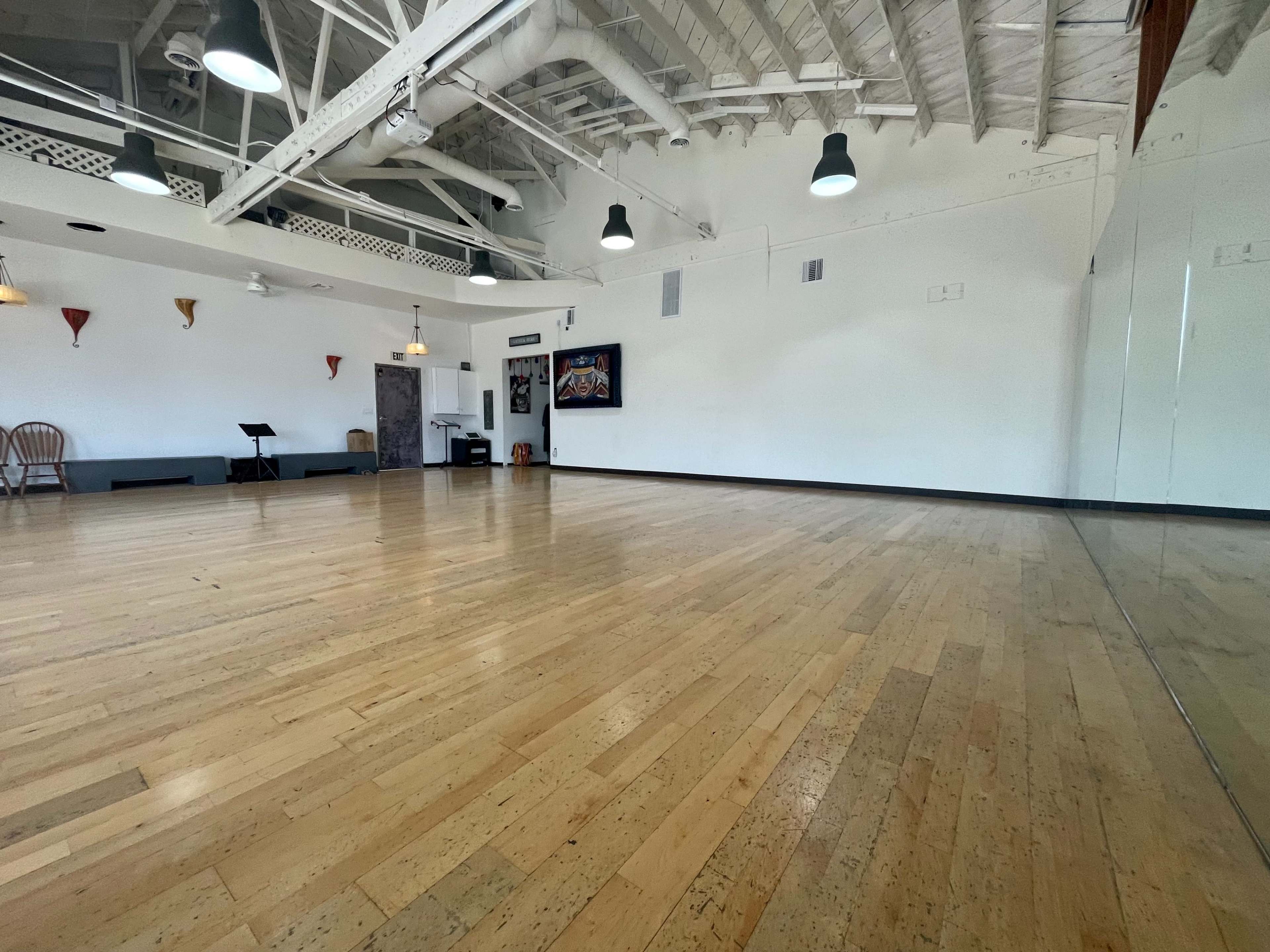 The image shows a spacious dance studio with polished wooden floors, white walls, and a large mirror along one side.