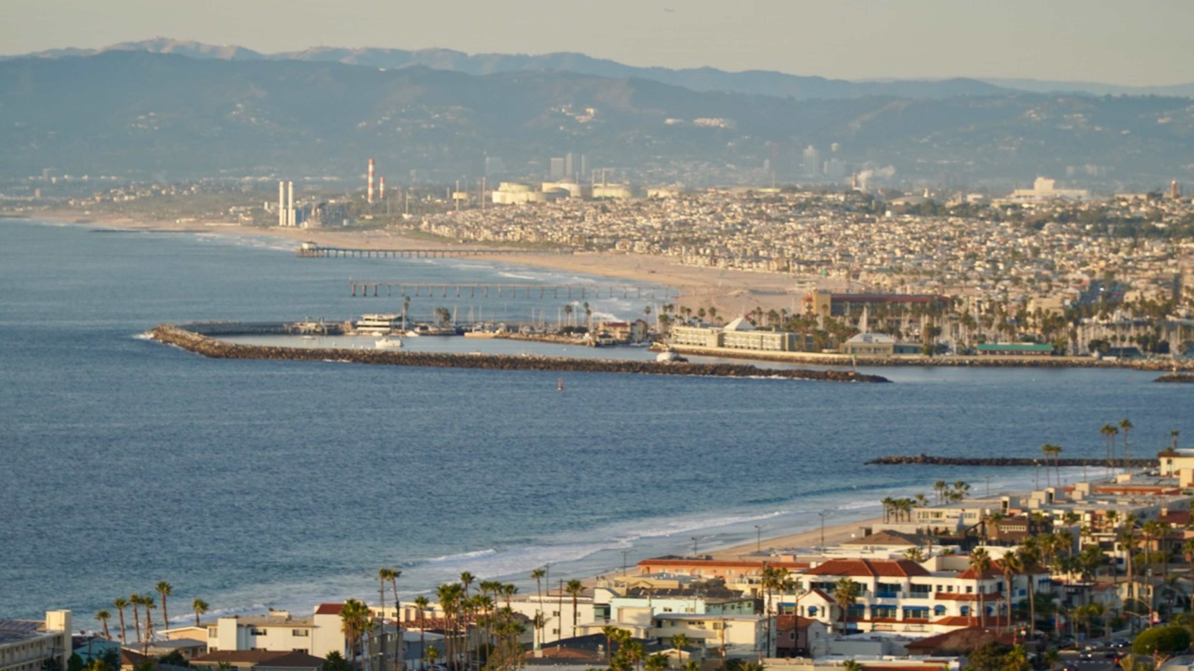 The image shows a coastal view featuring a city skyline, a pier, and a beach lined with buildings and palm trees.