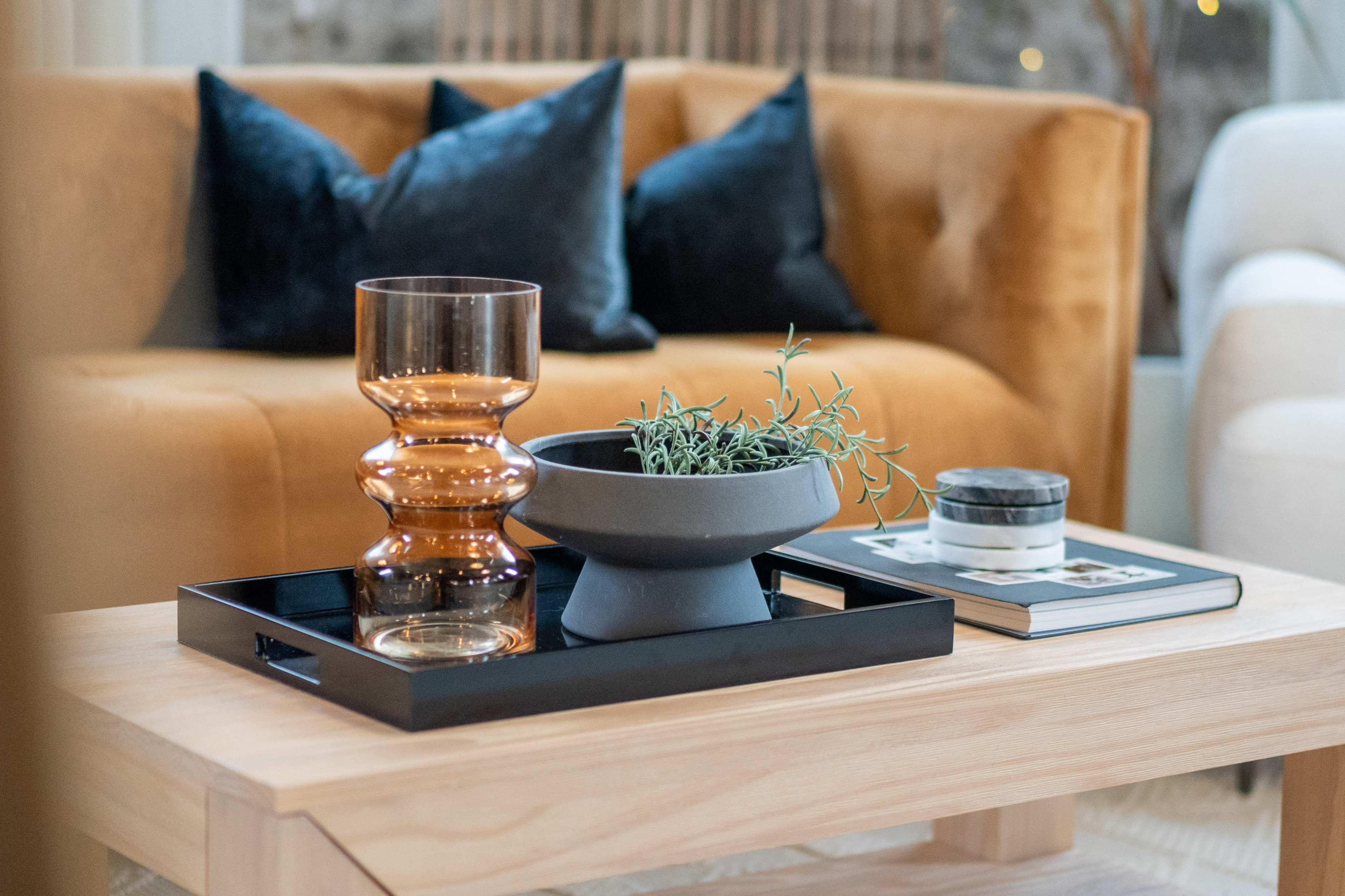 A wooden coffee table holds a decorative tray with a glass vase, a gray planter with greenery, and a small stack of magazines beside a plush couch with dark cushions.