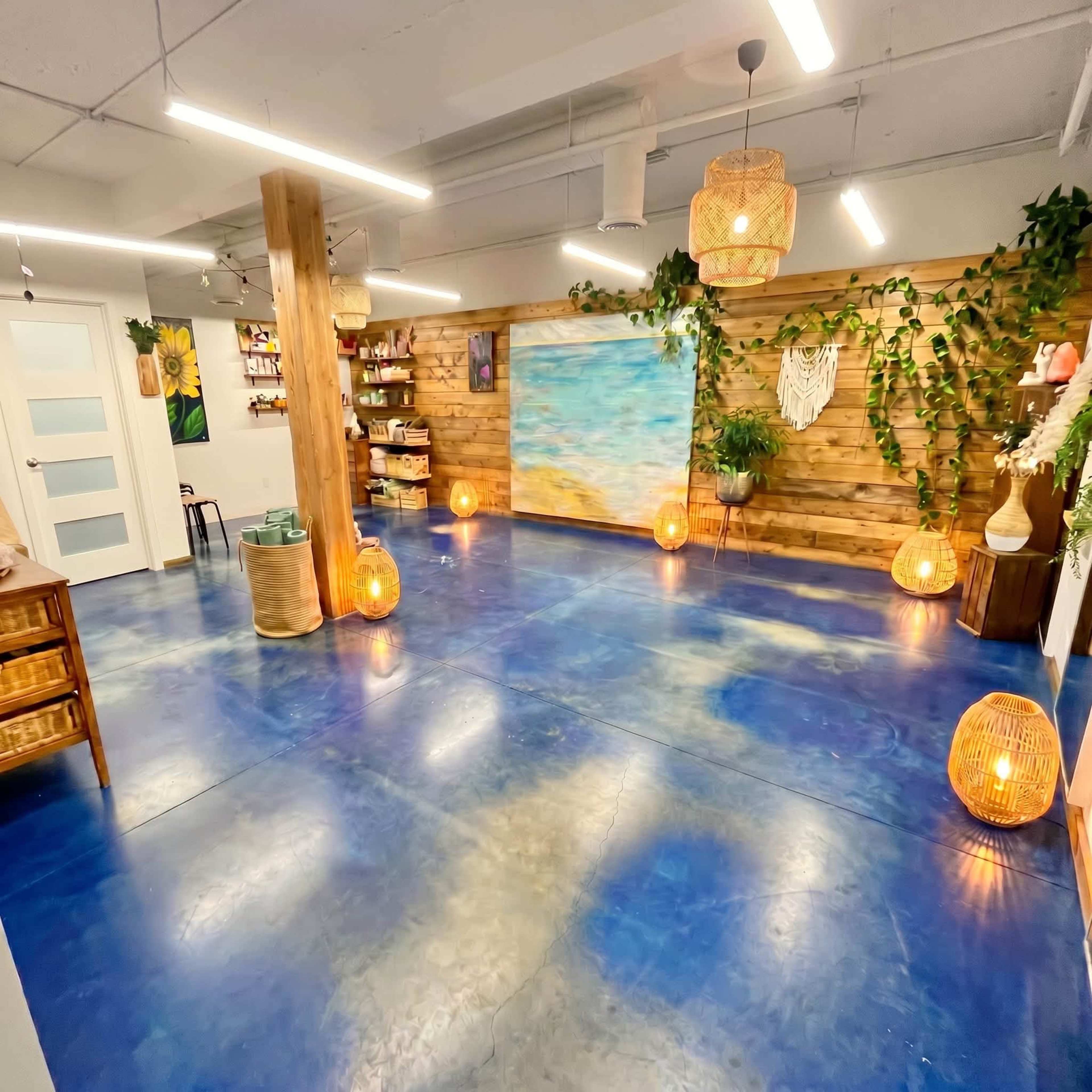 The image shows a brightly lit room with a blue floor, wooden walls, decorative plants, and hanging light fixtures, featuring a large painting of a beach scene on one wall.