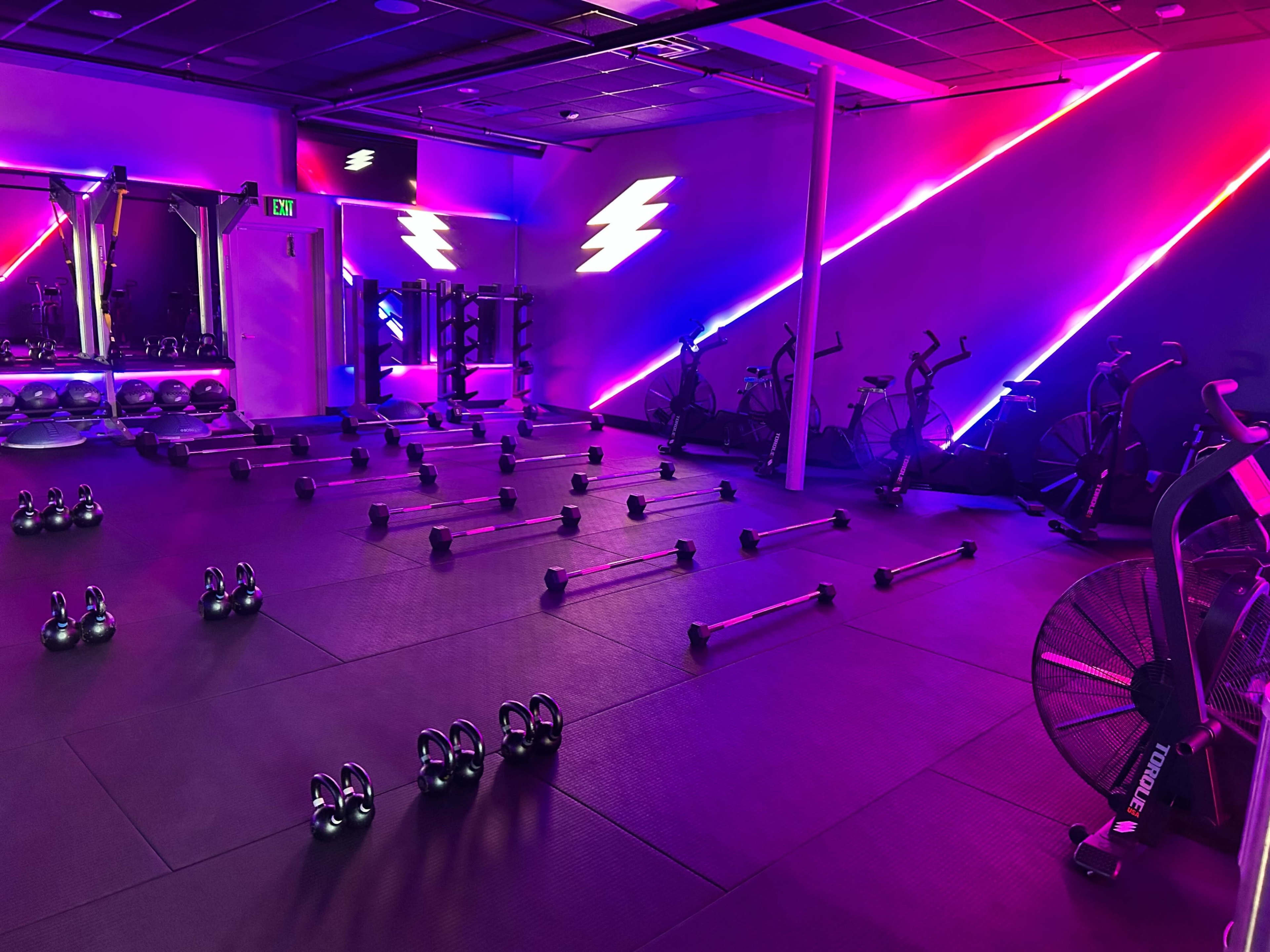 The image shows a fitness studio with an array of kettlebells and barbells arranged on the floor, stationary bikes lined against the wall, and colorful neon lighting illuminating the space.