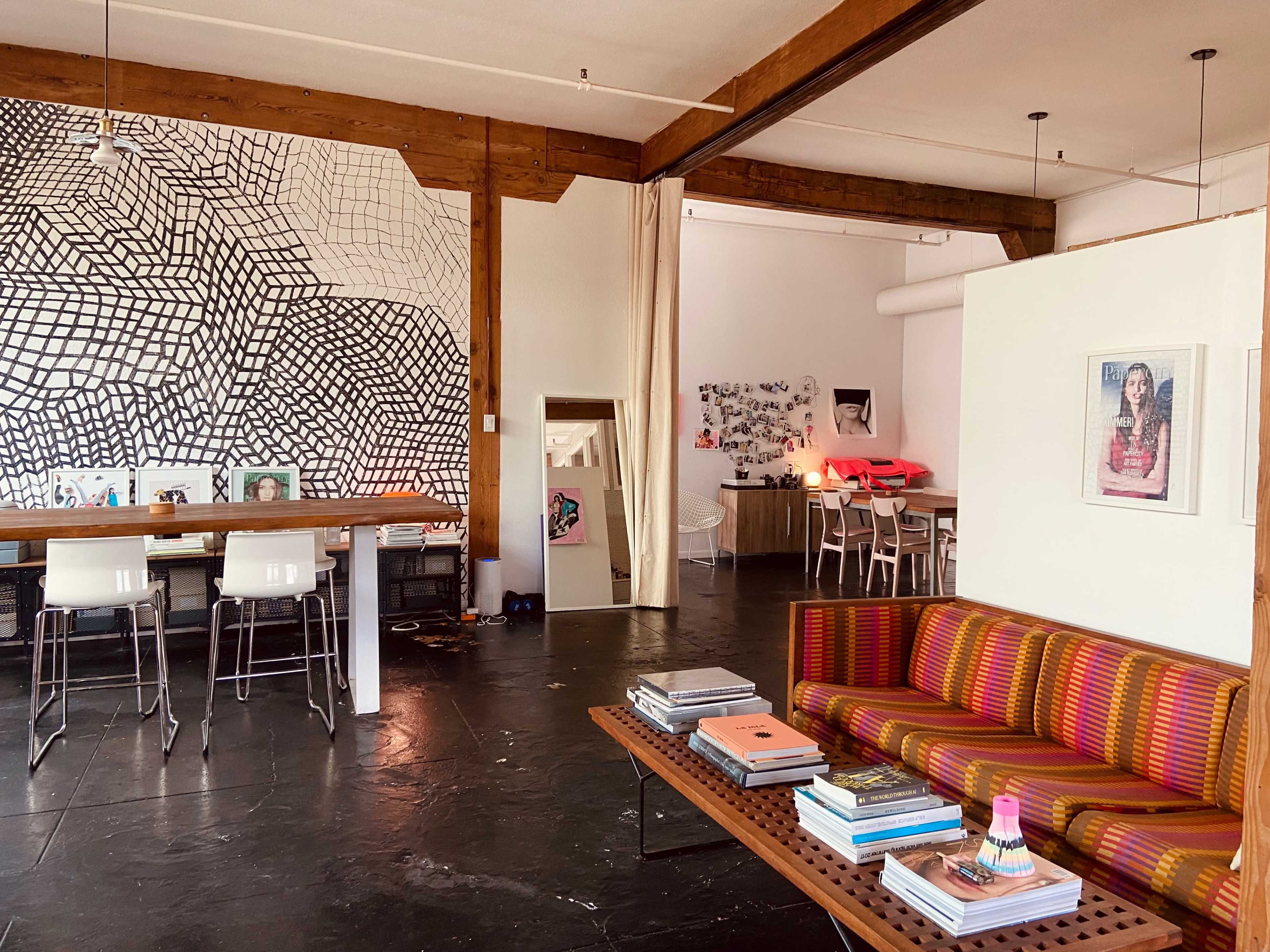 The image depicts a modern, open-concept living space featuring a colorful striped sofa, a long wooden table with stools, and a wall adorned with a geometric mural.