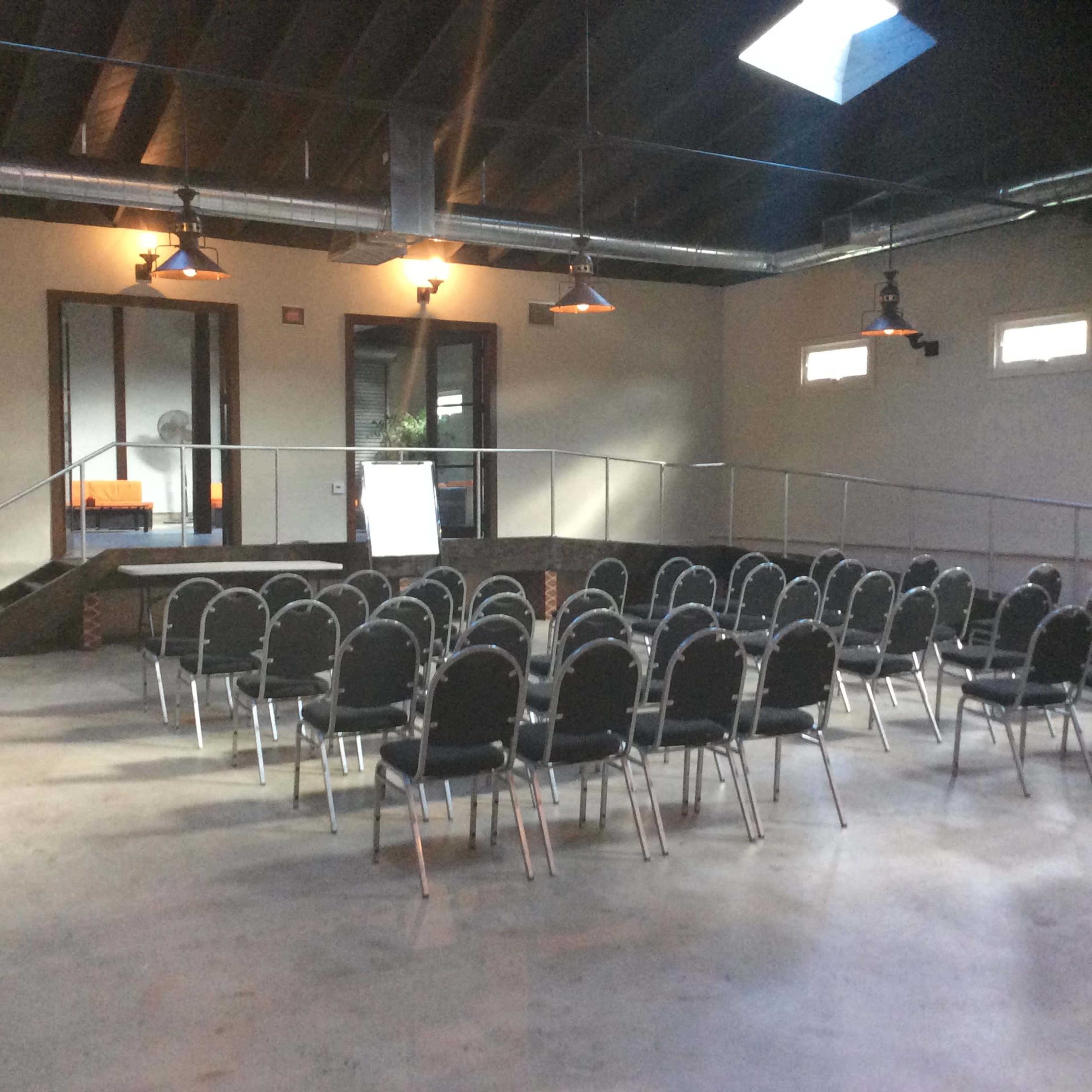 The image shows a spacious room set up with rows of black chairs facing a small stage area, illuminated by overhead lights.