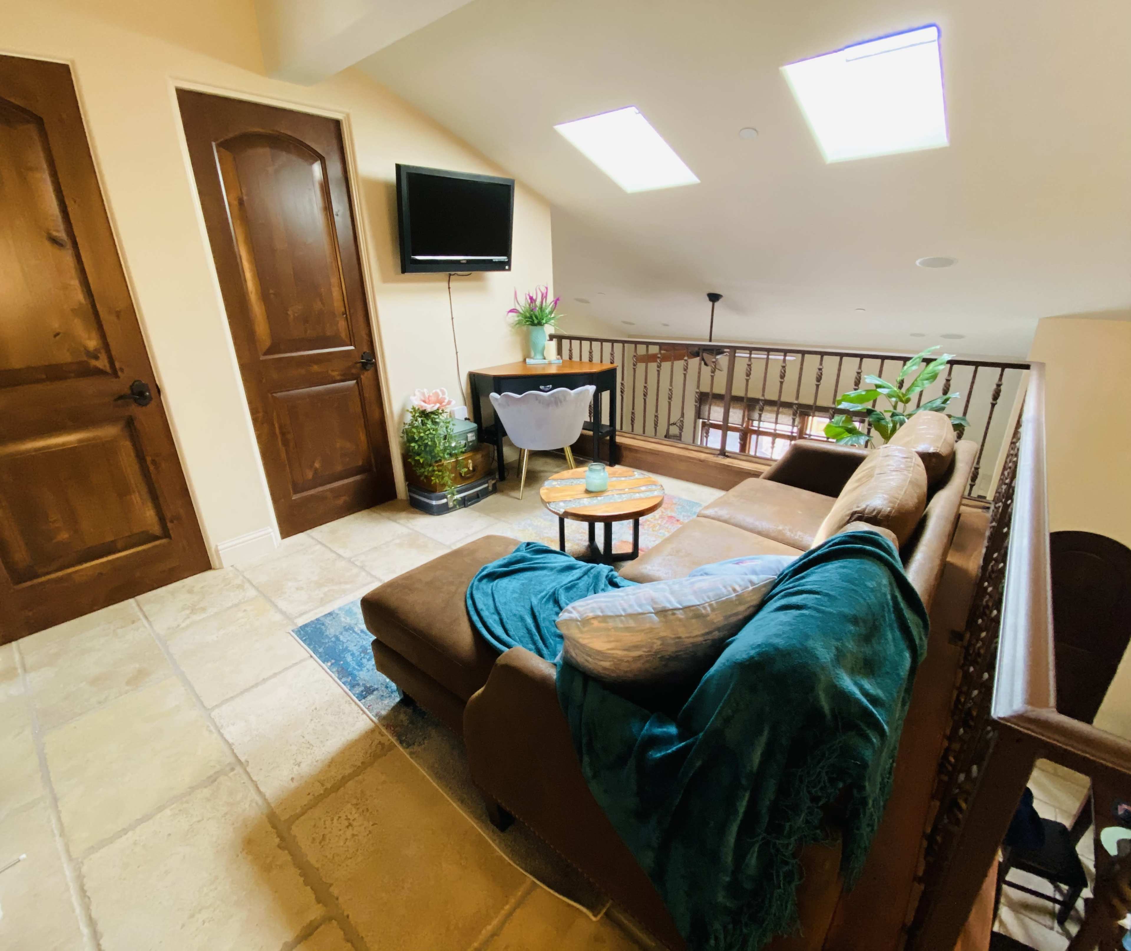 The image shows a cozy loft area with a brown sectional sofa, a small coffee table, and a television mounted on the wall, illuminated by natural light from skylights.