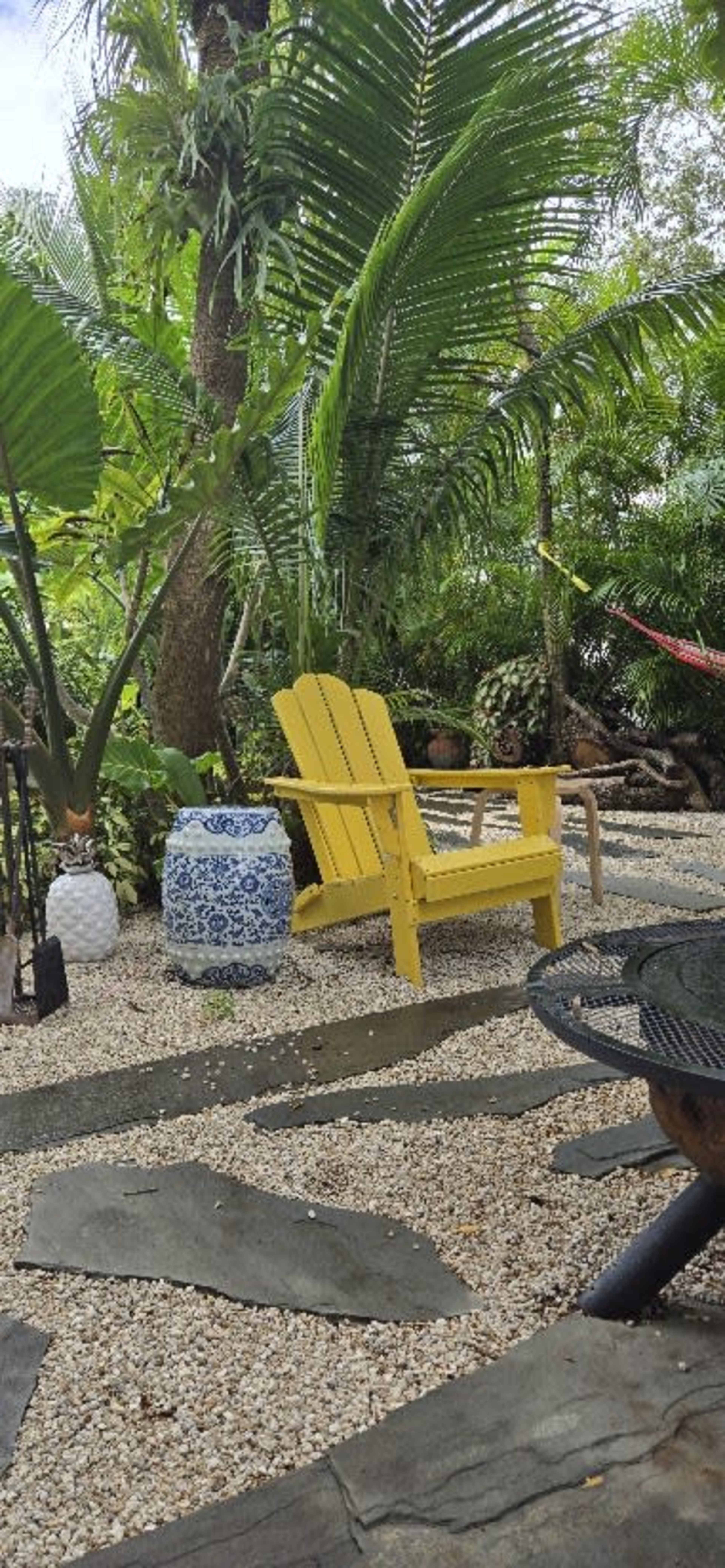A yellow Adirondack chair is set among tropical plants and stone pathways in a garden.