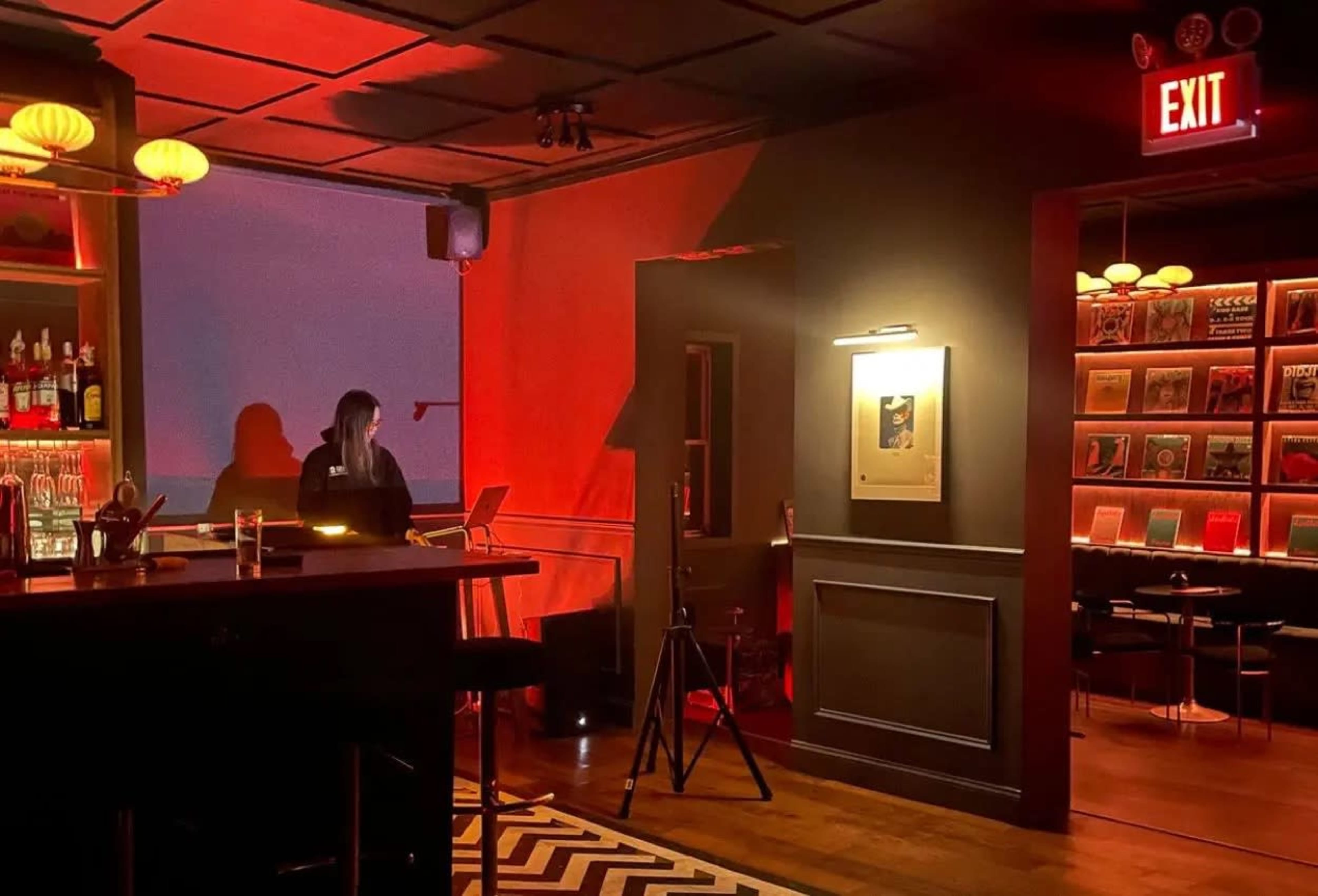 A dimly lit bar scene features a woman at a counter, with vibrant red lighting and an exit sign visible in the background.