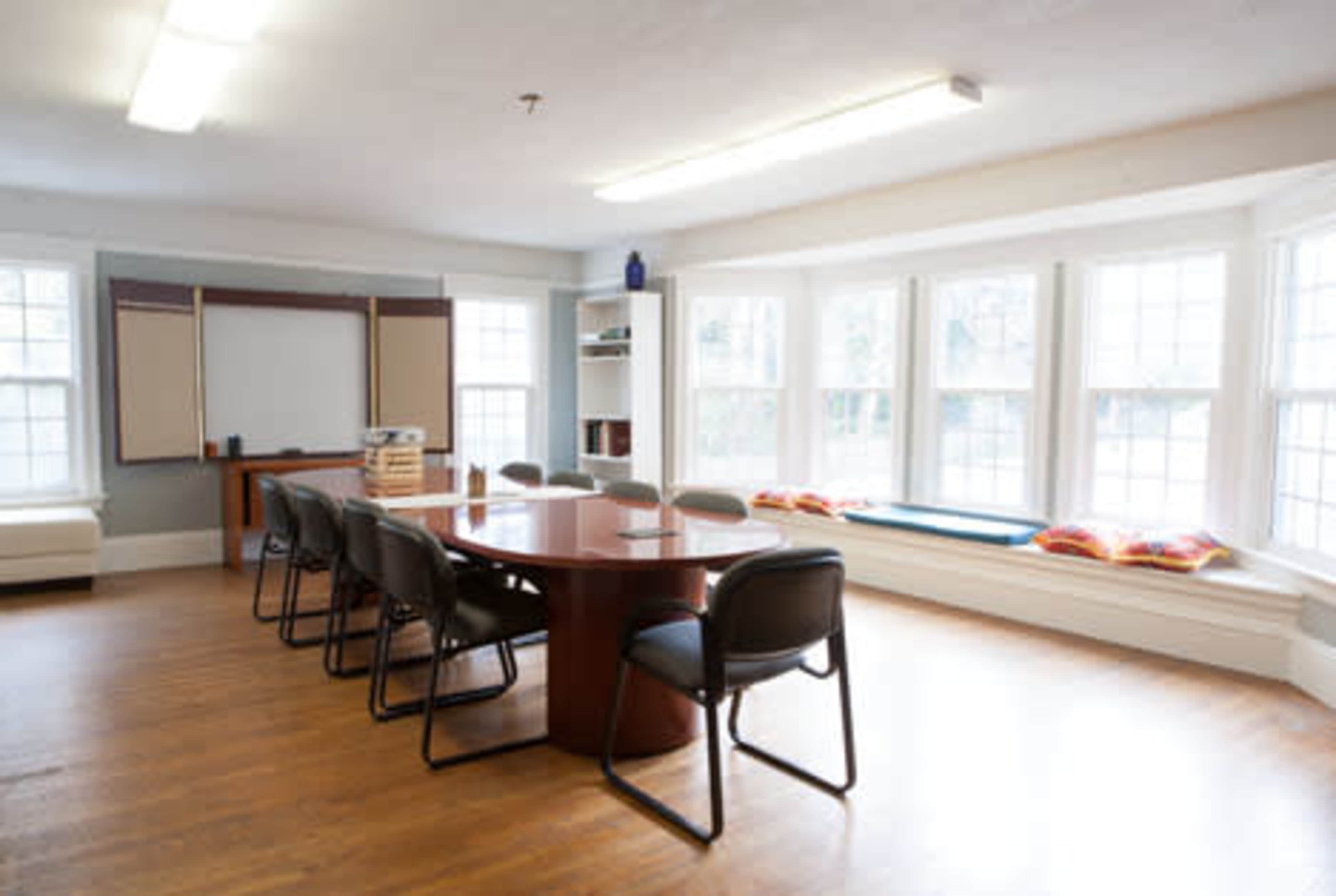 The image shows a conference room with a large round table surrounded by several chairs, a whiteboard, and large windows with seating.