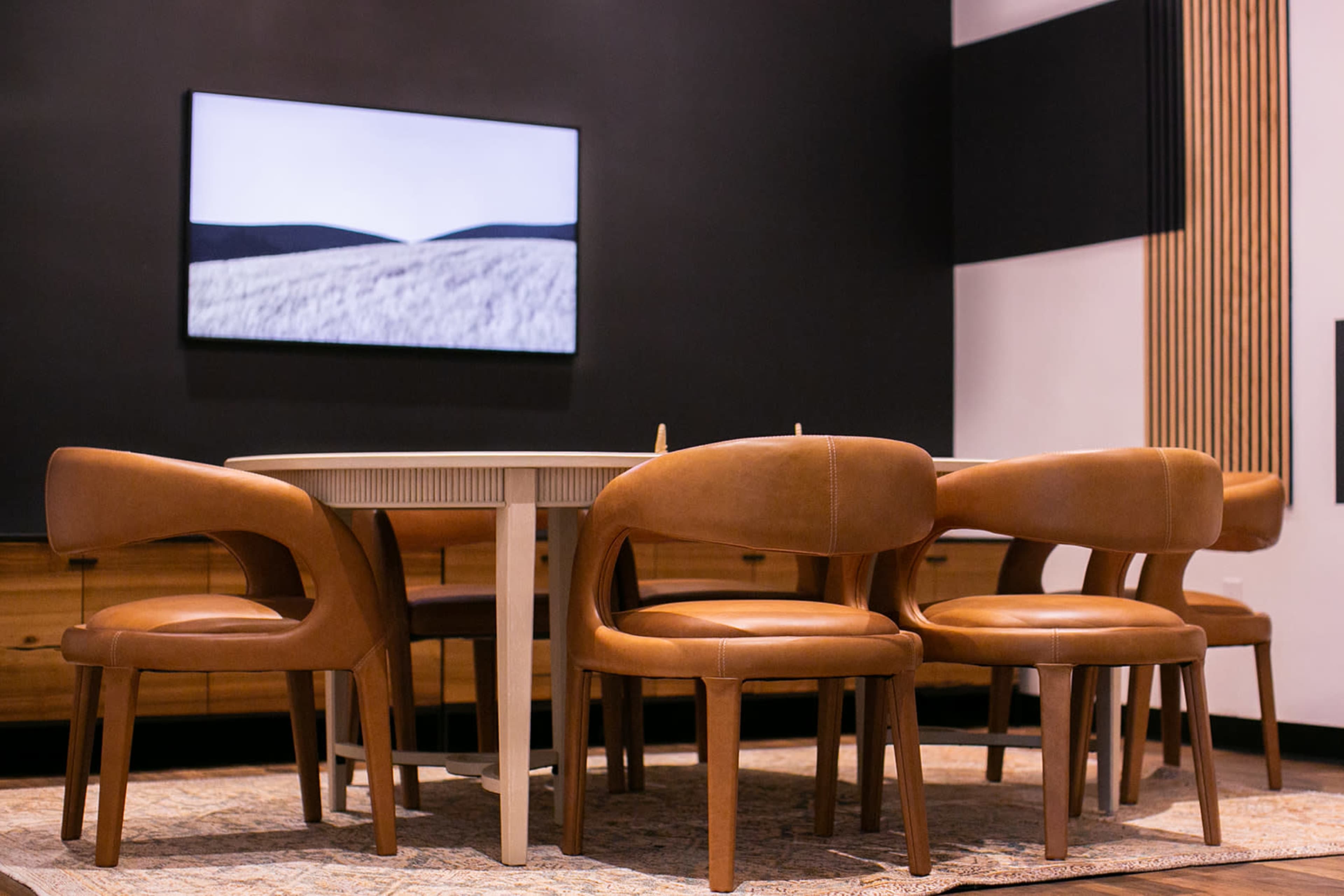A round dining table is surrounded by five brown upholstered chairs in a modern room with a dark accent wall and a wall-mounted television displaying a black-and-white landscape.