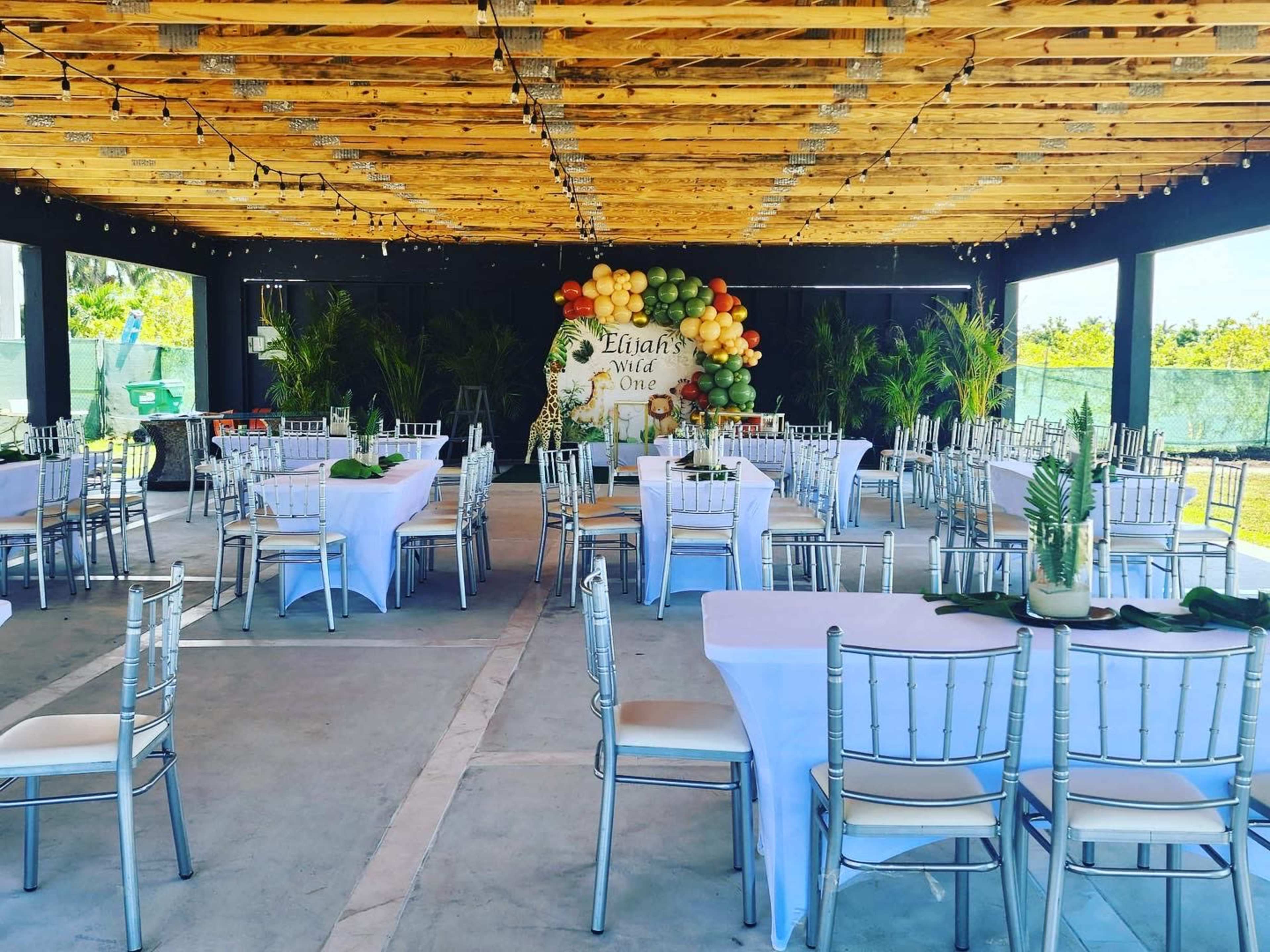 The image shows an event space with multiple tables and chairs arranged under a wooden ceiling, decorated with a colorful balloon backdrop.