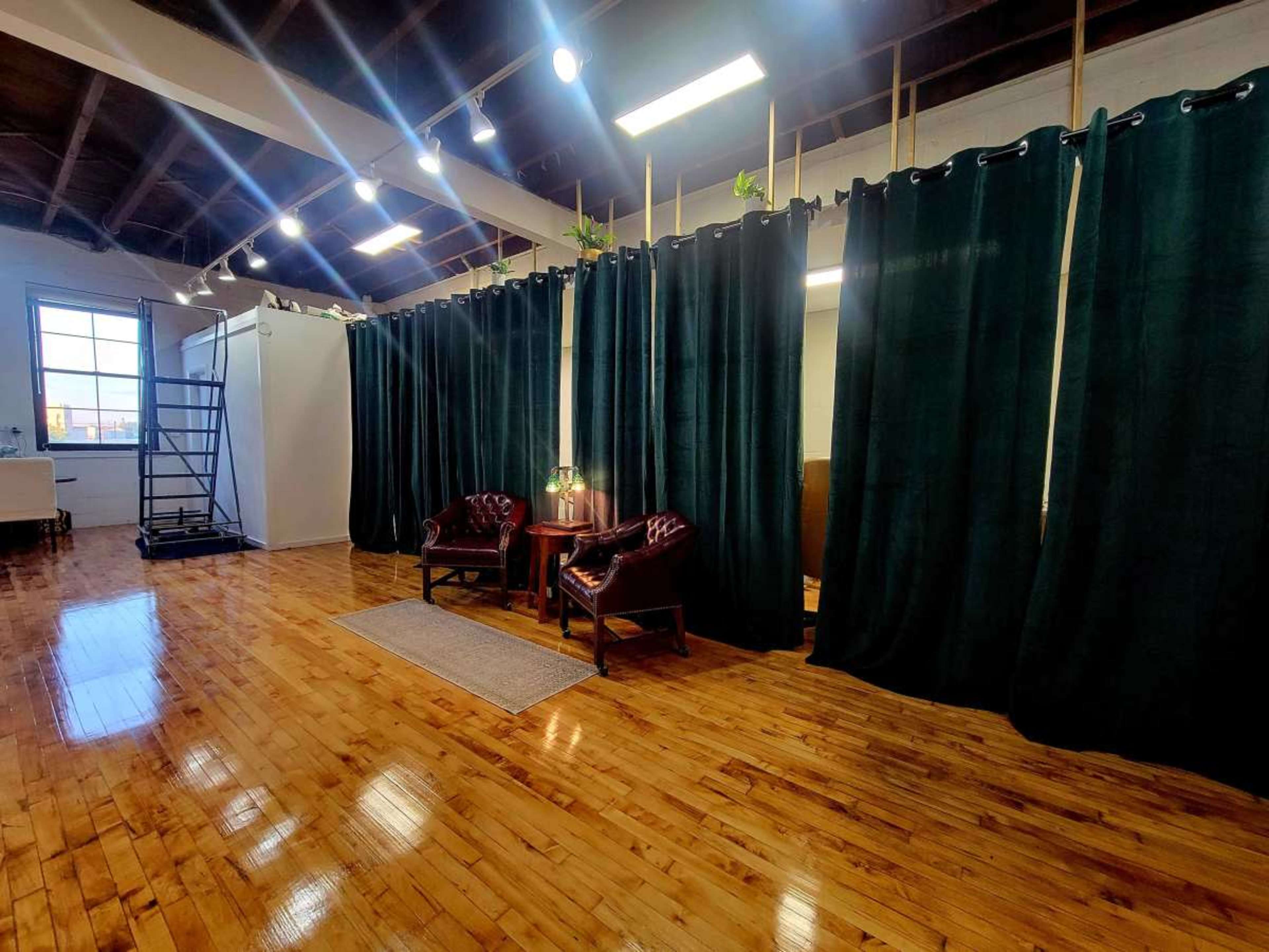 The image shows a spacious room with wooden floors, featuring two leather chairs, a small side table, and dark green curtains dividing the space.