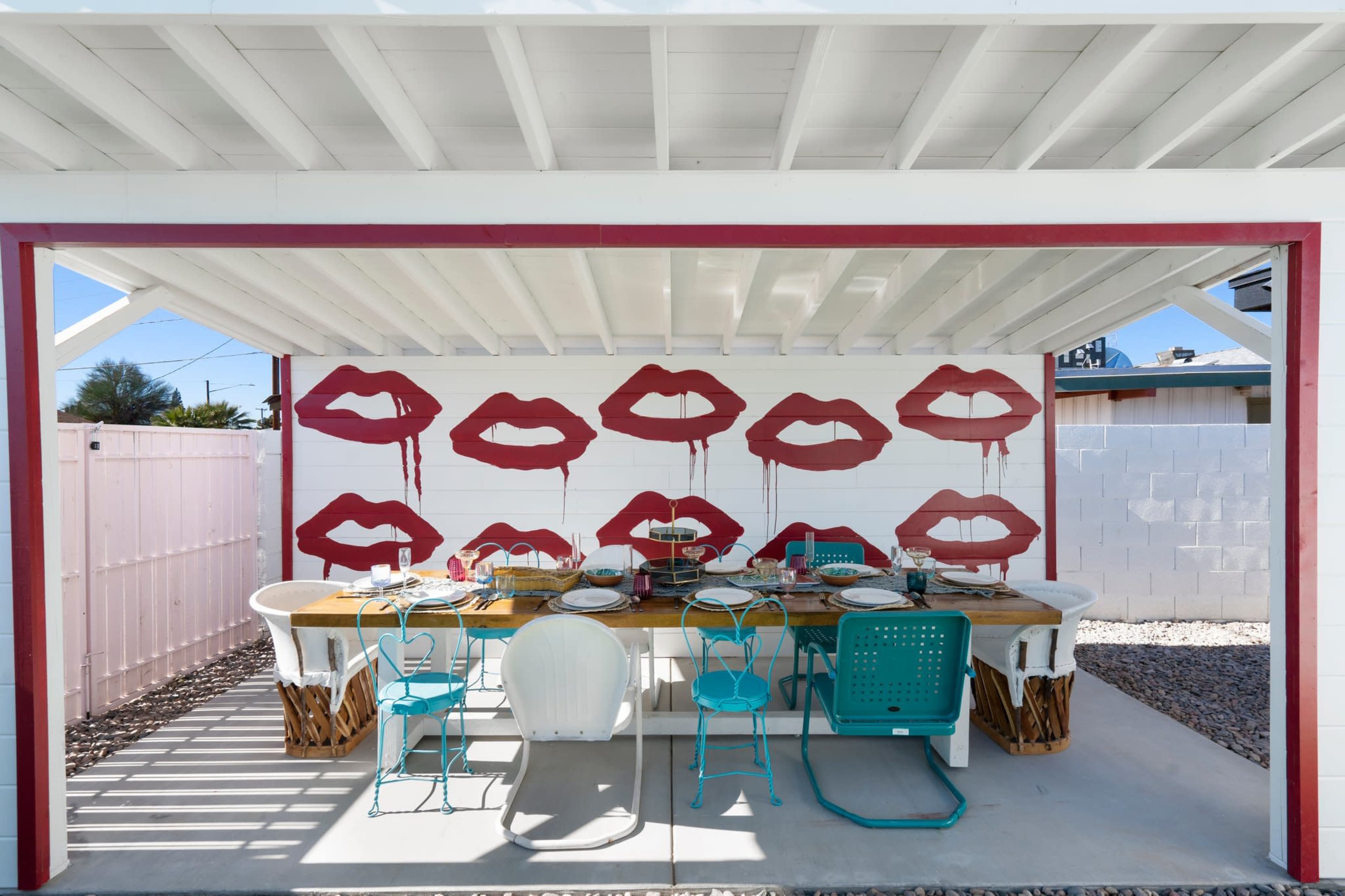 The image shows an outdoor dining area under a covered patio with a large table set for a meal and decorated with red lip prints on the walls.