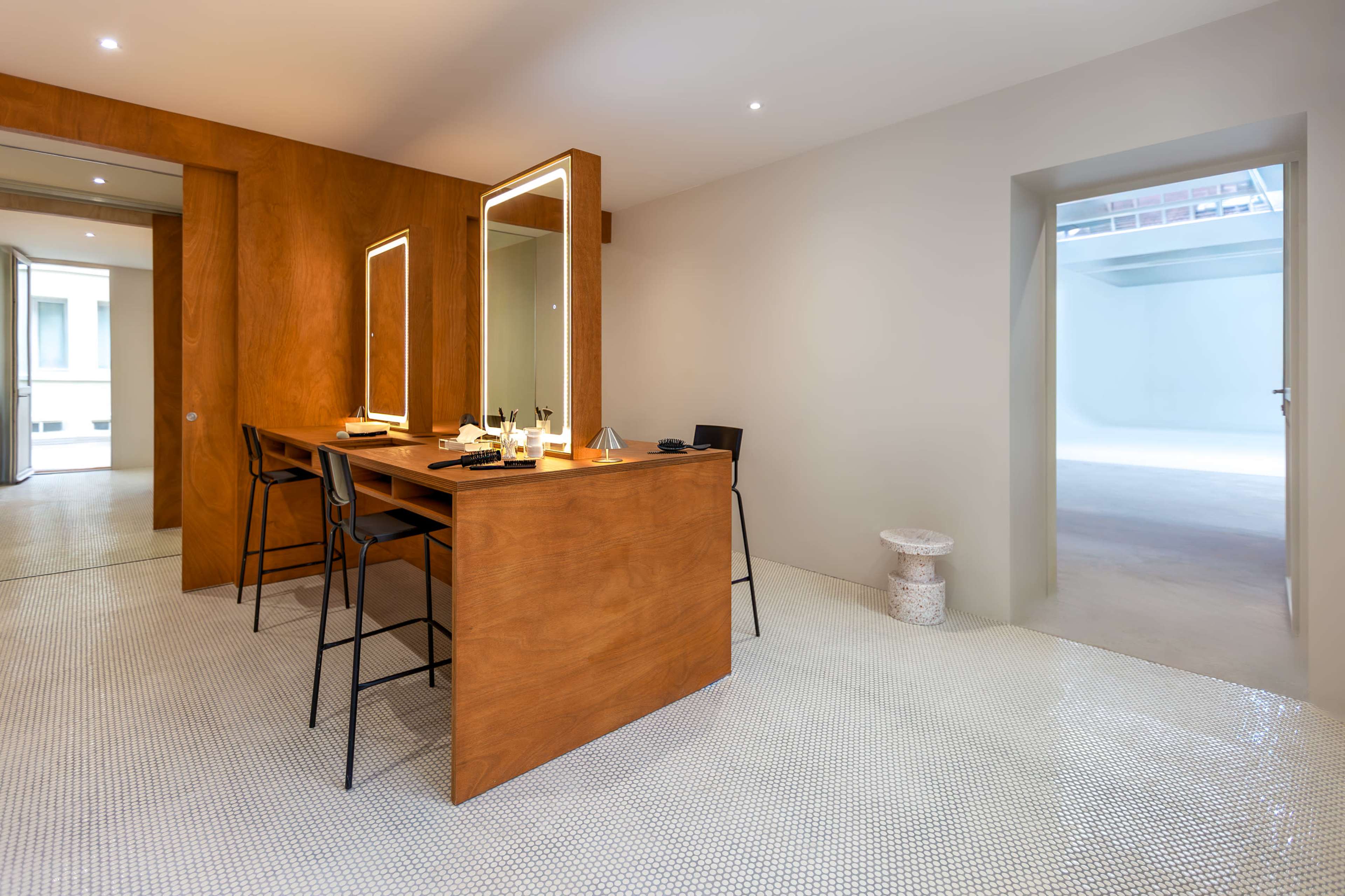The image shows a modern, minimalist makeup vanity area with wooden panels, large mirrors, and a well-lit open space.