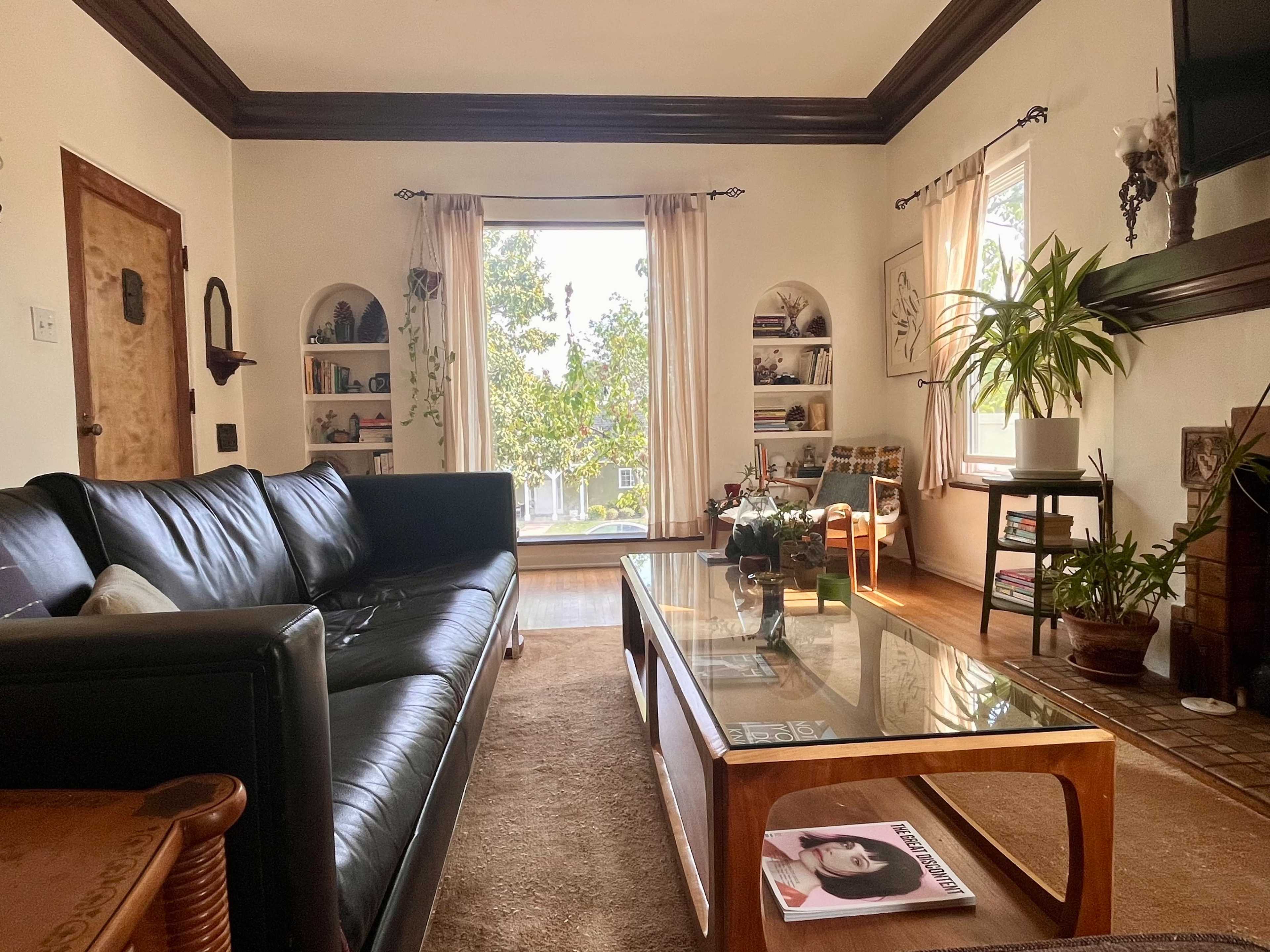 The living room features a black leather couch, a wooden coffee table with magazines, and large windows allowing natural light to illuminate the space.