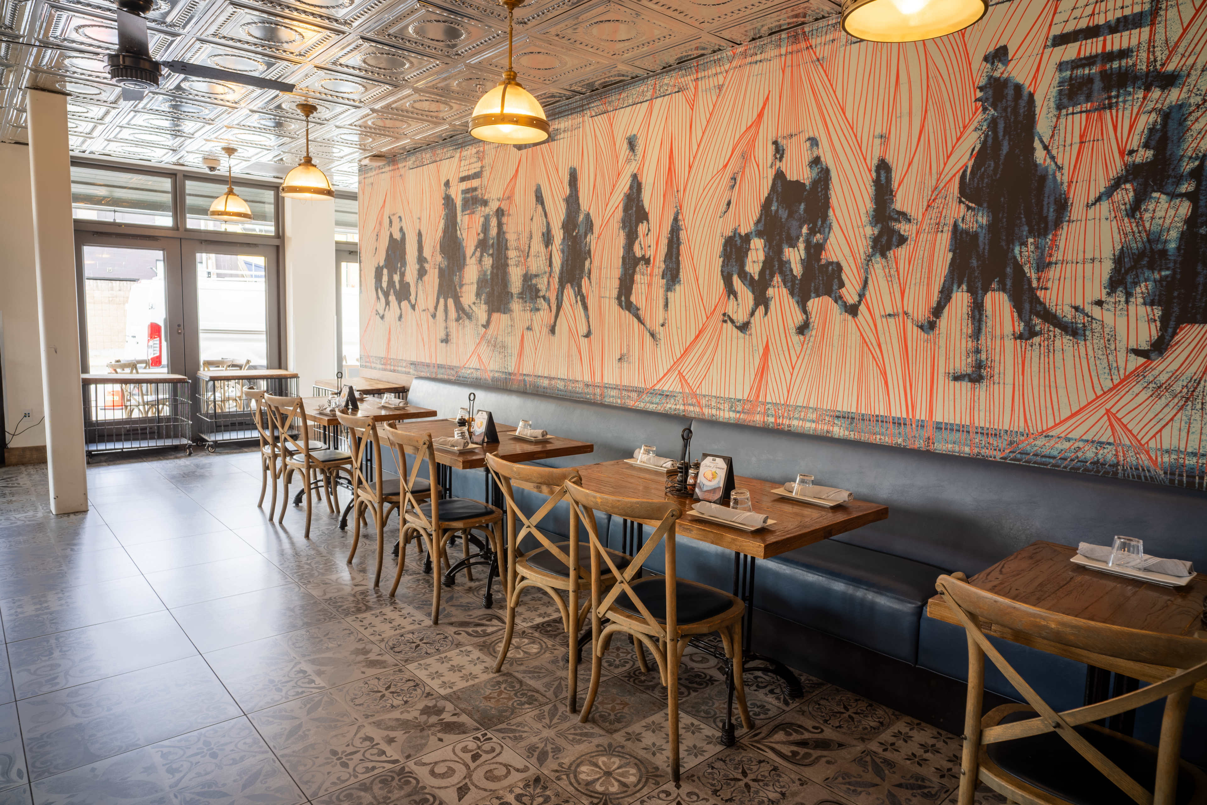 The image shows an interior of a restaurant featuring wooden tables, cross-back chairs, and a large mural of people walking along one wall.