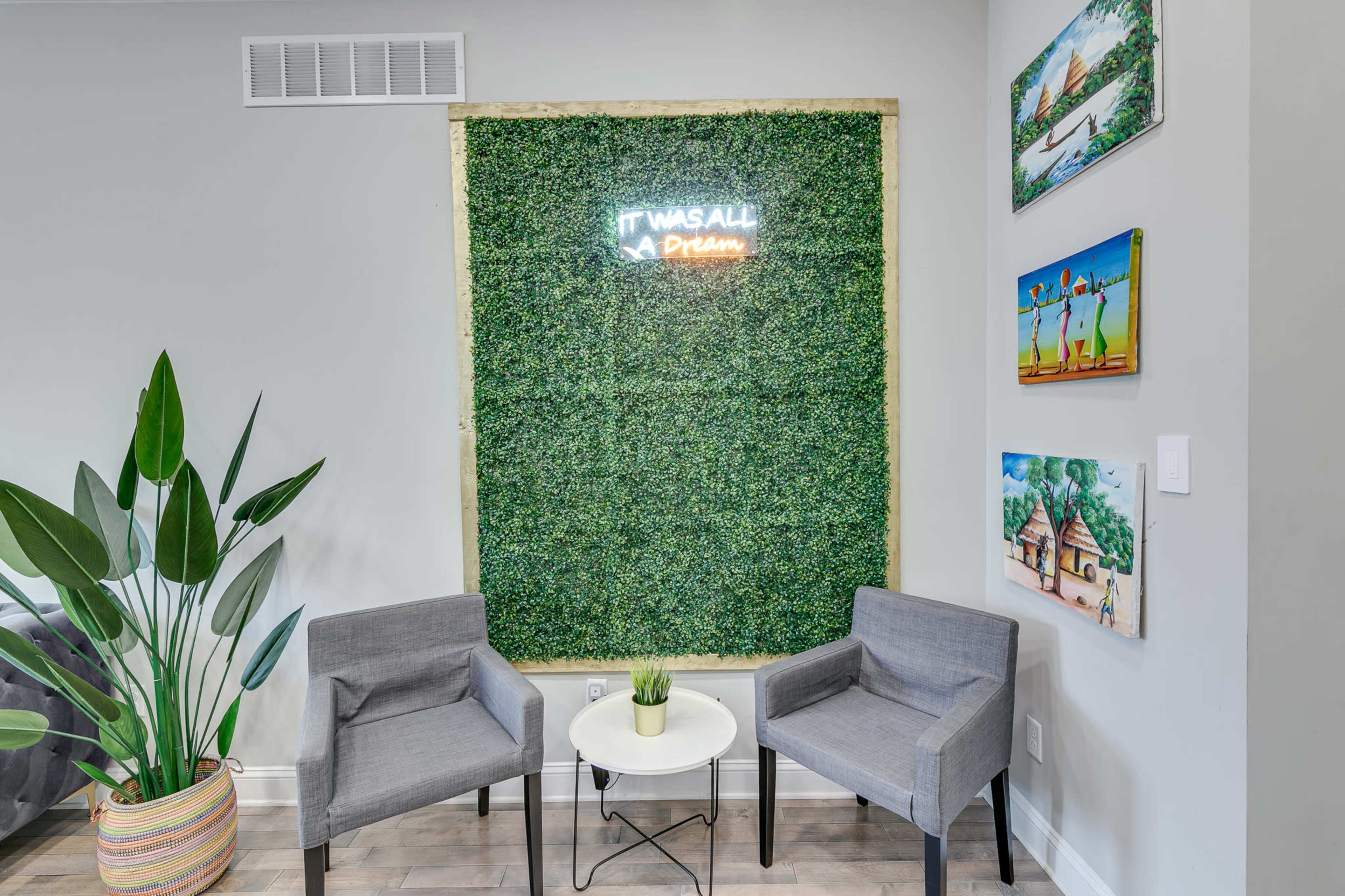 A cozy indoor space featuring two gray chairs facing a small white table, surrounded by a green wall and decorated with colorful artwork.