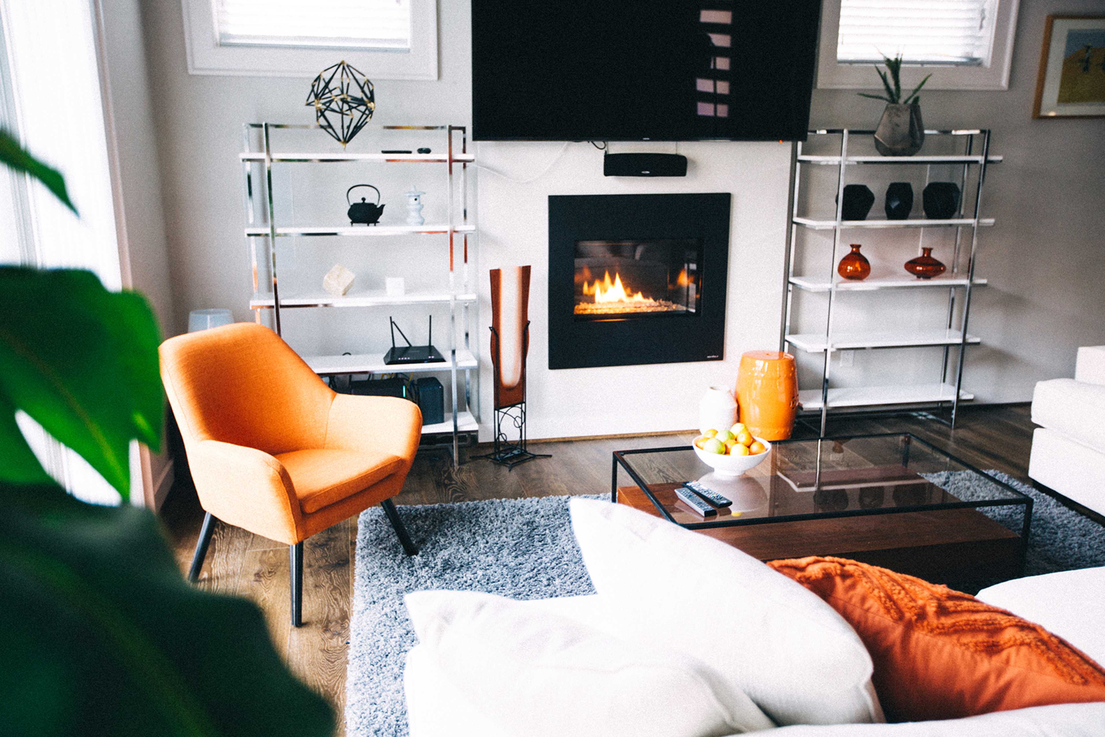 A modern living room features an orange chair, a glass coffee table, and a fireplace with decorative shelves displaying vases and sculptures.