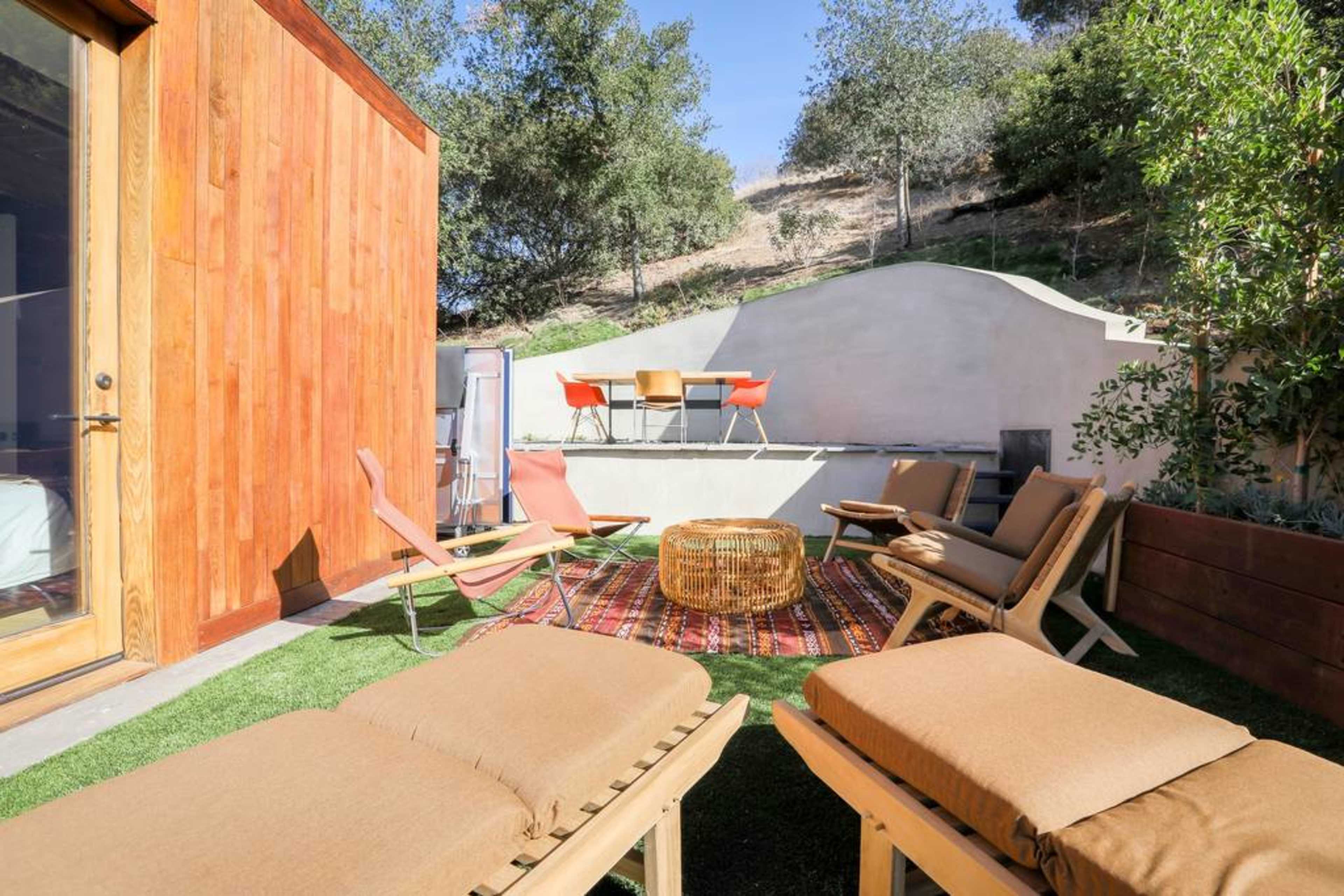 The image shows a backyard space with wooden seating, a round wicker table, and a colorful rug surrounded by greenery and hills.