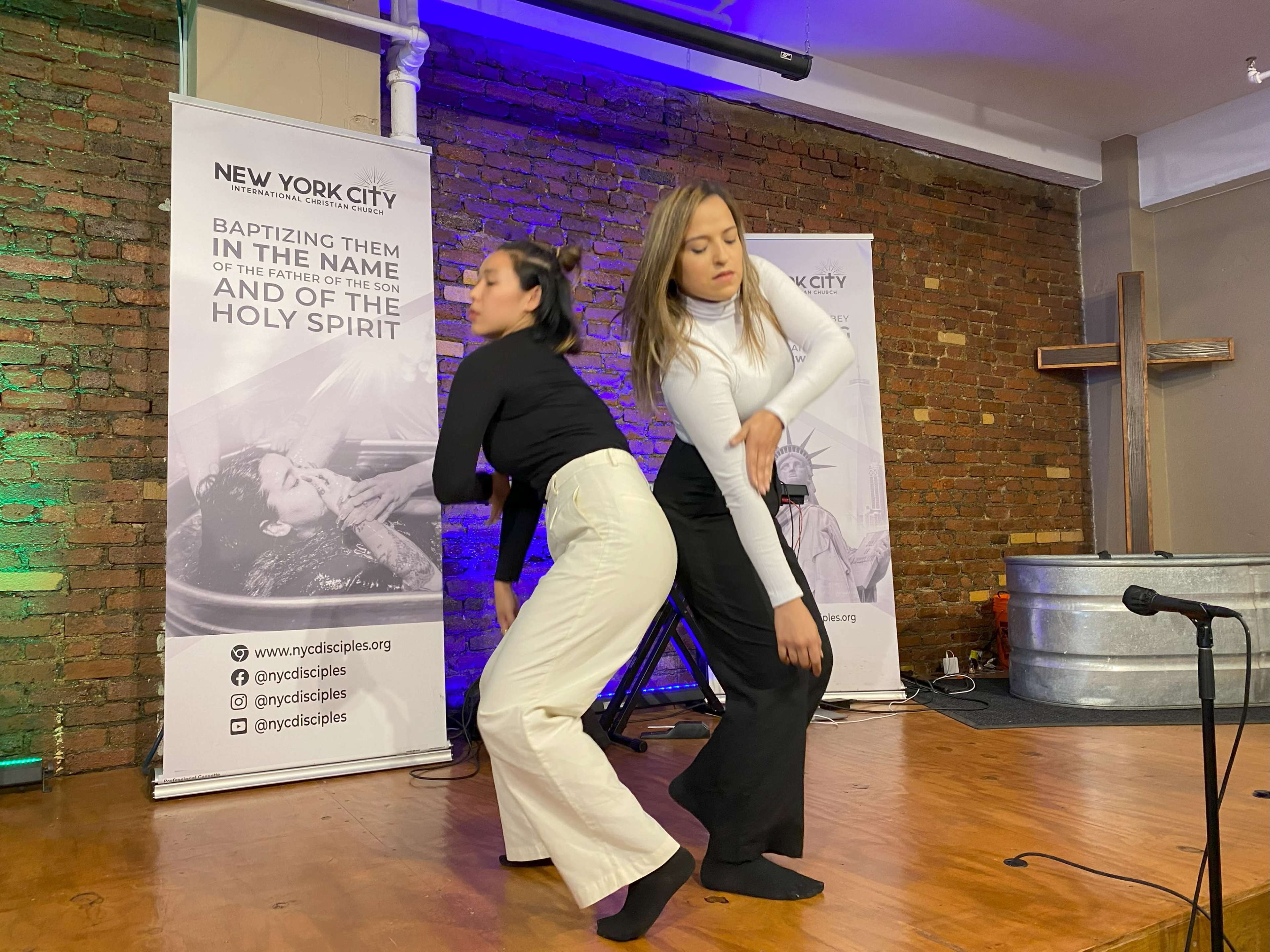 Two women perform a dance in front of a backdrop featuring banners and a wooden cross in a well-lit venue.