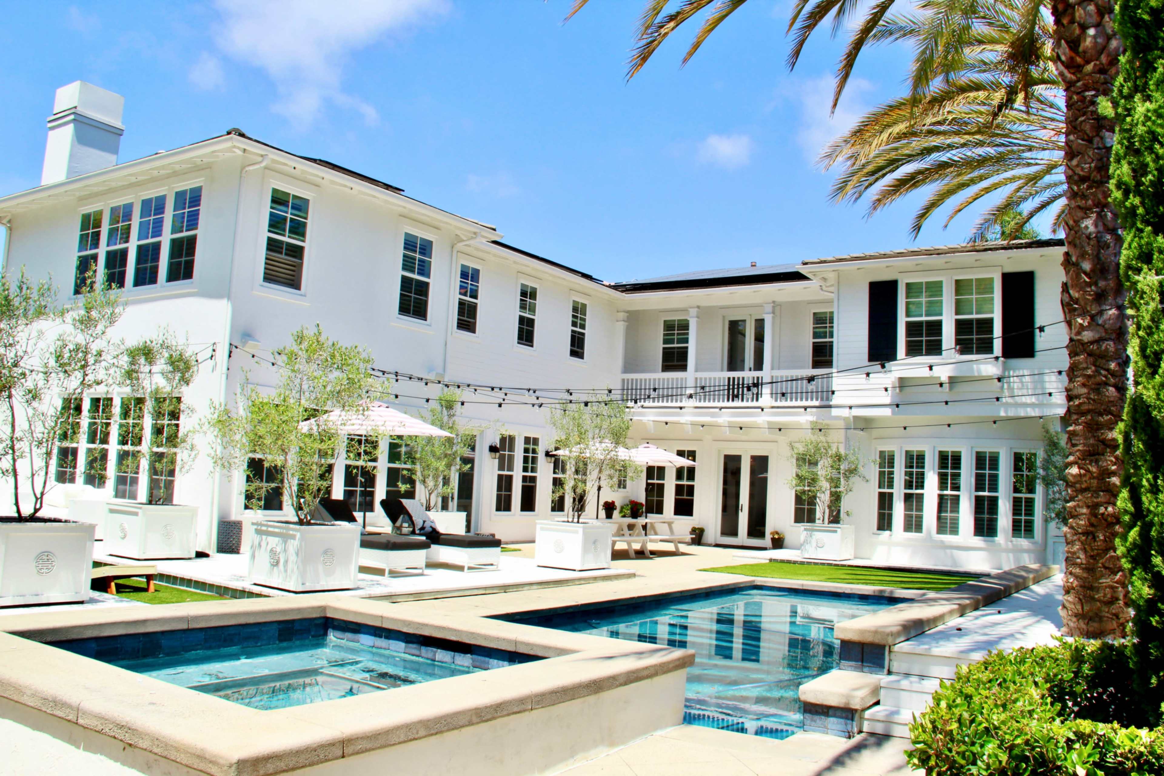 The image shows a modern white house with multiple balconies, surrounded by palm trees and featuring two swimming pools in the courtyard.