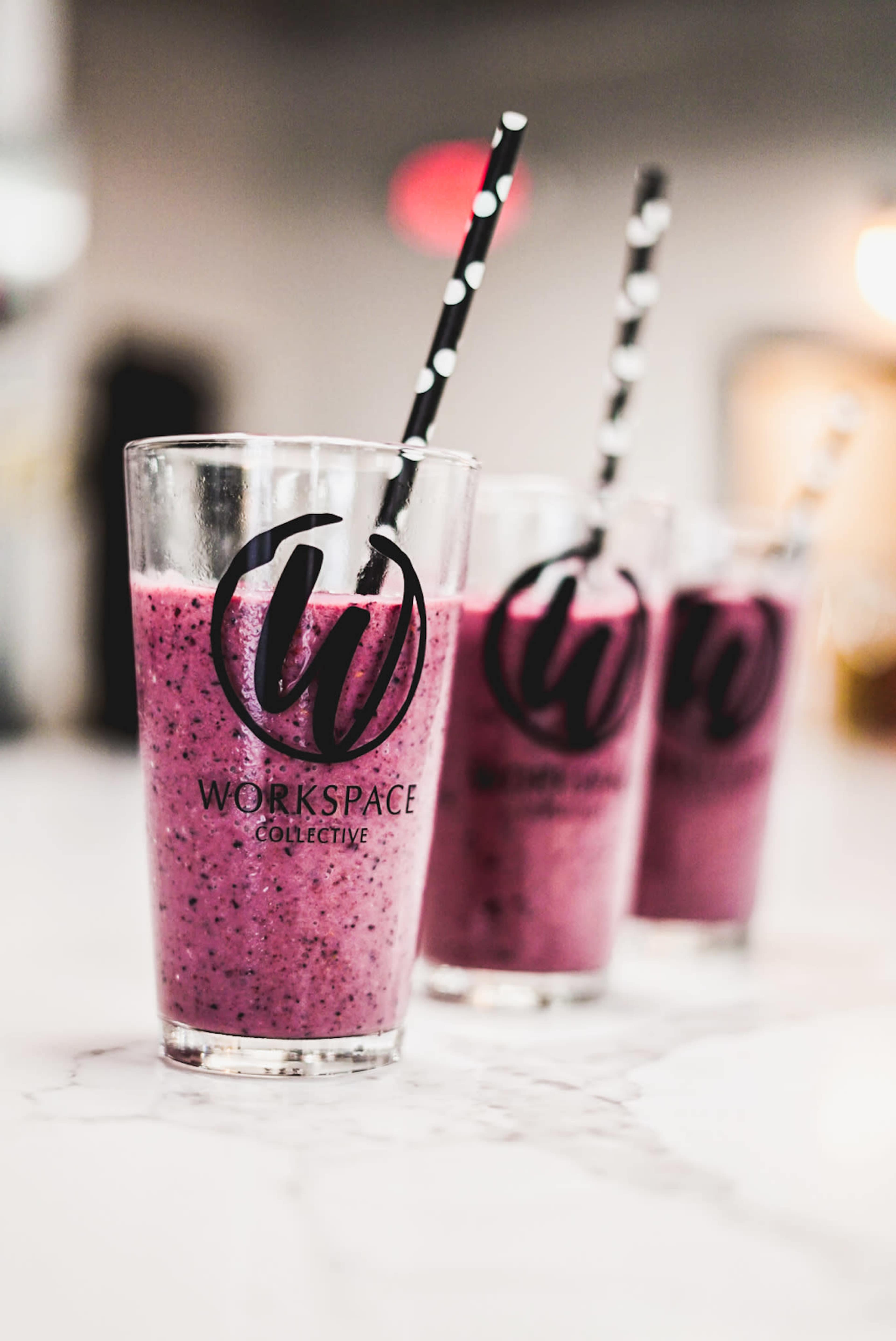 Three clear glasses containing purple smoothies with black and white striped straws are lined up on a marble countertop, each featuring a logo that reads "Workspace Collective."