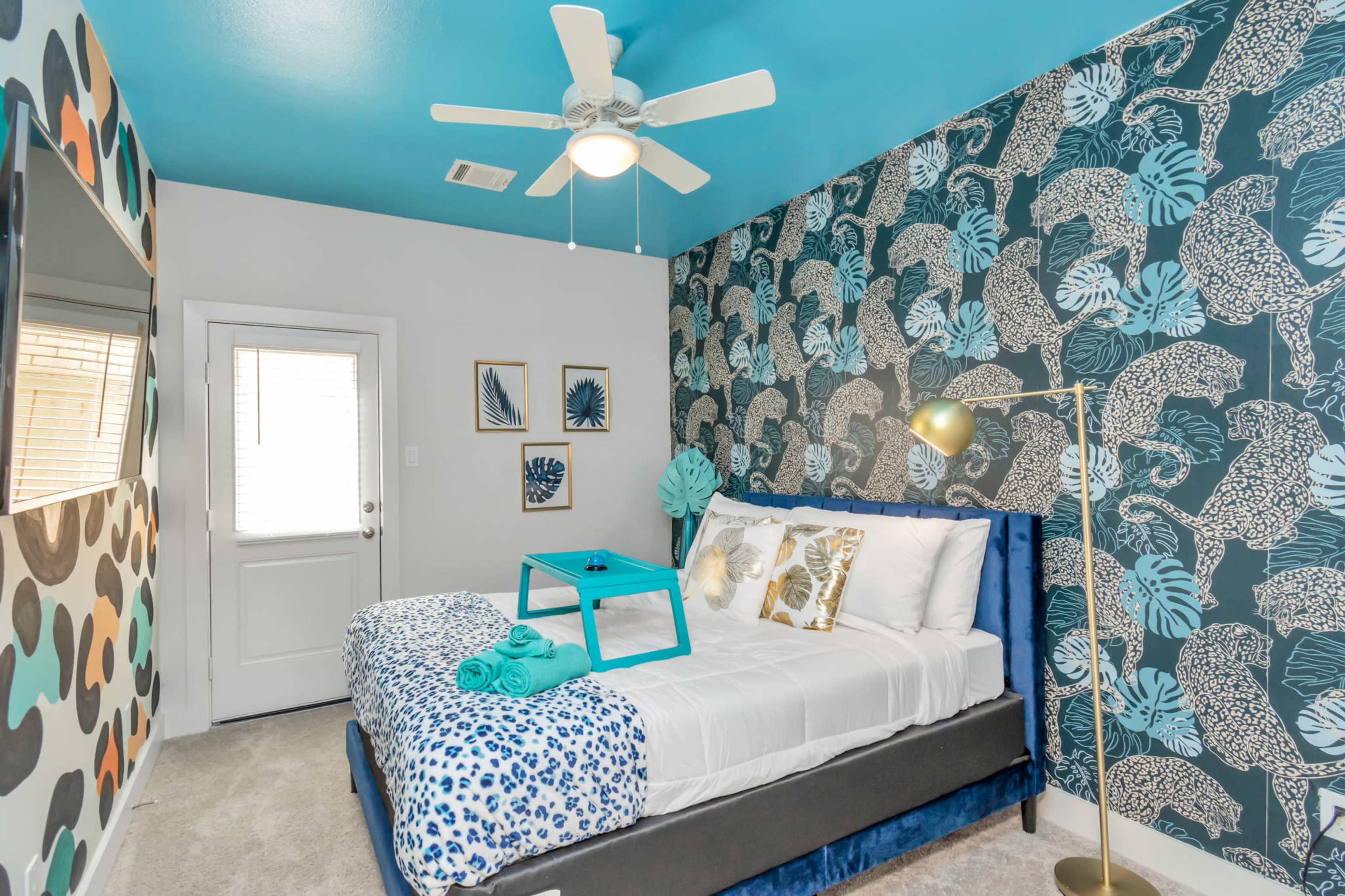 A modern bedroom featuring a bed with a blue and white patterned blanket, a teal side table, a gold floor lamp, and a wall decorated with a colorful floral pattern.