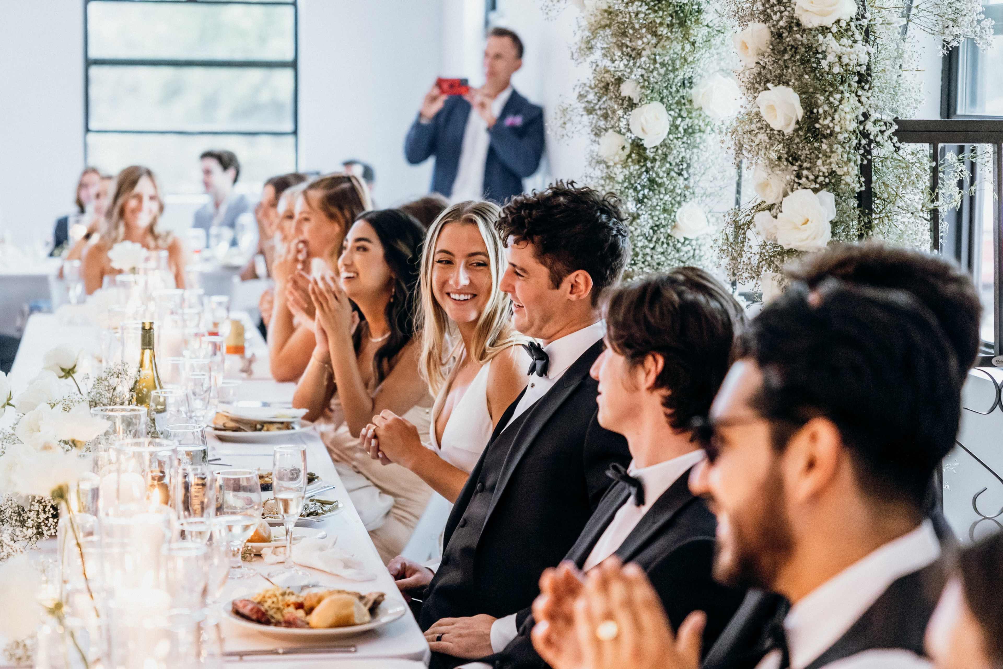 A group of elegantly dressed guests sits at a long table, smiling and clapping during a wedding reception.