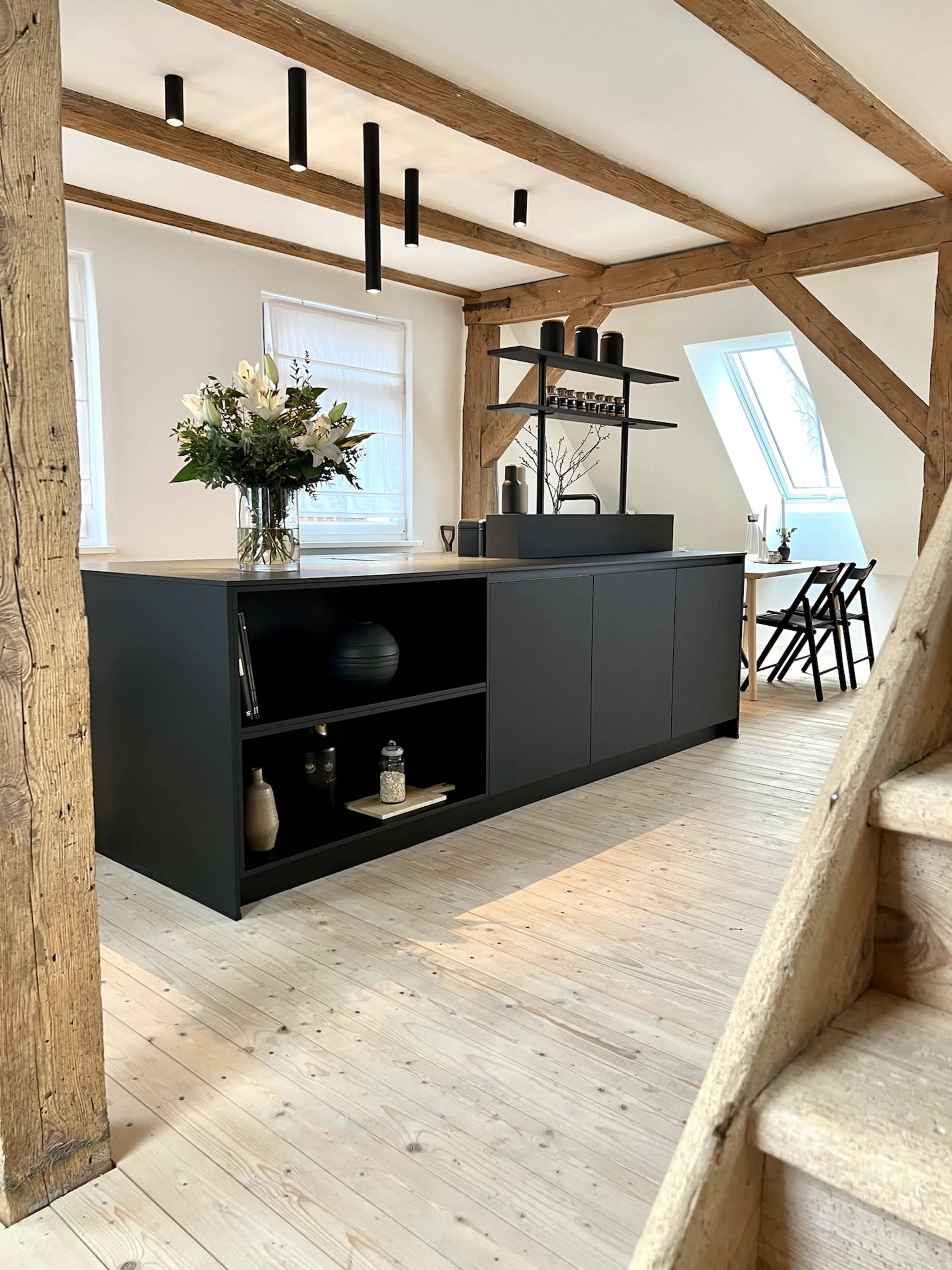 A modern kitchen features a central island with dark cabinetry, wooden beams, and a light wooden floor.
