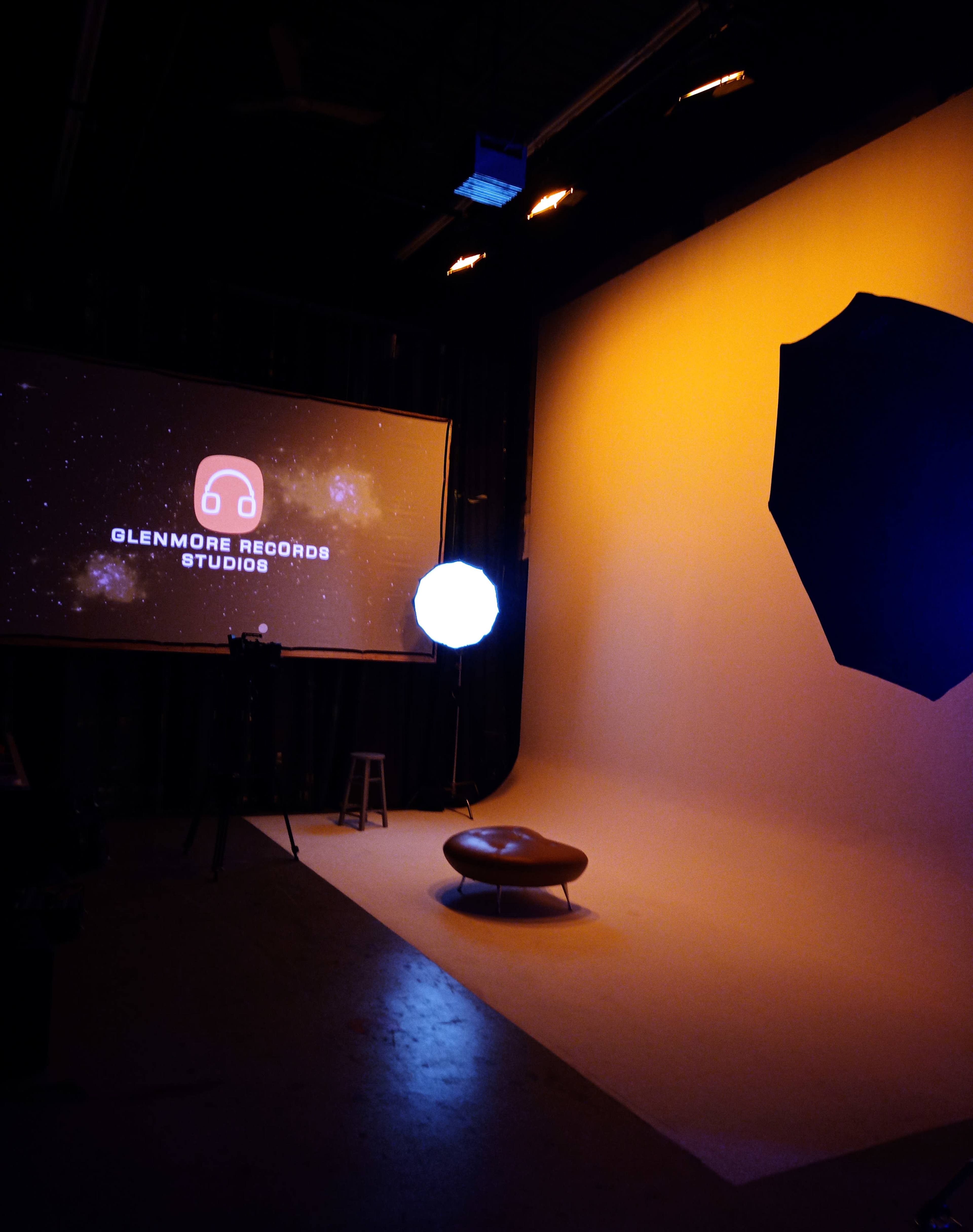 The image shows a studio setup with a large screen displaying "Glenmore Records Studios," a stool, and lighting equipment in a sparsely furnished space.